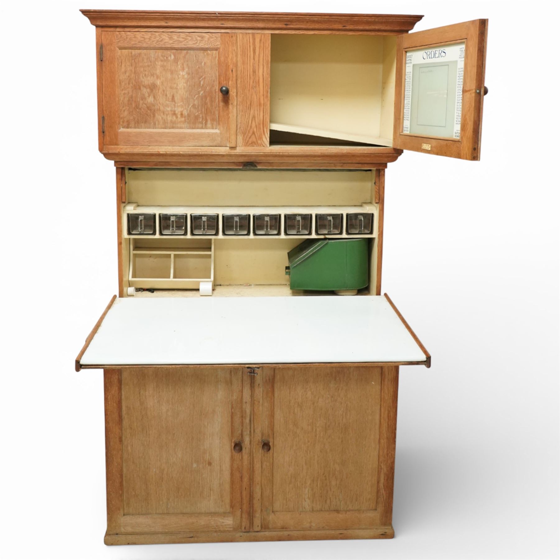 Early 20th century 'Quicksey' oak kitchen larder, rectangular moulded cornice over a pair of panelled cupboard doors, the fall front enclosing a white enamel work surface with fitted interior comprising original glass drawers, storage compartments and shelf, the lower section with twin panelled cupboard doors enclosing shelving and divided storage, raised on a plinth base