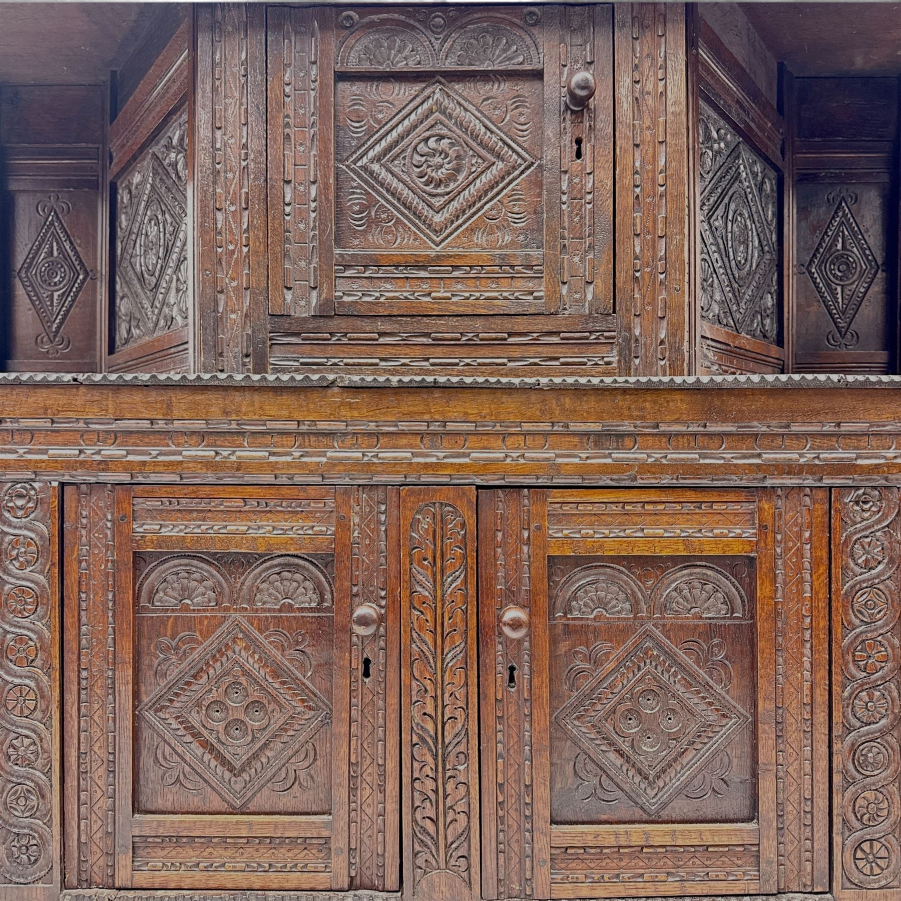 Early 20th-century  oak Jacobean two-tier revival court cupboard, rectangular top with carved notch and stylized foliate carved edge above a carved lozenge pattern frieze and a central cupboard with carved geometric designs and geometric carved panelled sides, above a carved panelled cupboard with two doors of similar geometric design, the whole flanked by two sets of stop-fluted baluster turned columns and supported on stile feet