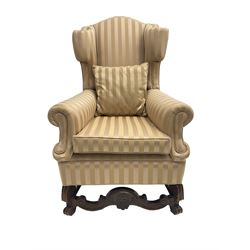 William and Mary style wingback armchair, upholstered in gold striped fabric, on walnut scroll carved supports joined by scroll and foliate carved middle rail, turned H-shaped stretchers, on castors
