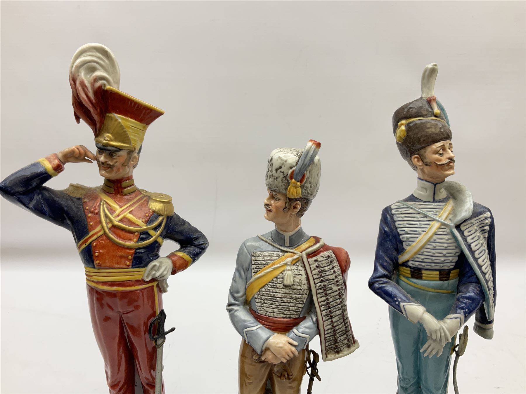 Group of seven Capodimonte figures of soldiers, to include four Bruno-Merli examples including Officer of the 12th Lancers 1820 and Captain of the 18th Hussars 1815, both raised upon ornate gilt plinths bearing British monarch motto in Latin 'Dieu et mon droit', and three further similar, all with crowned N mark beneath, tallest H32cm