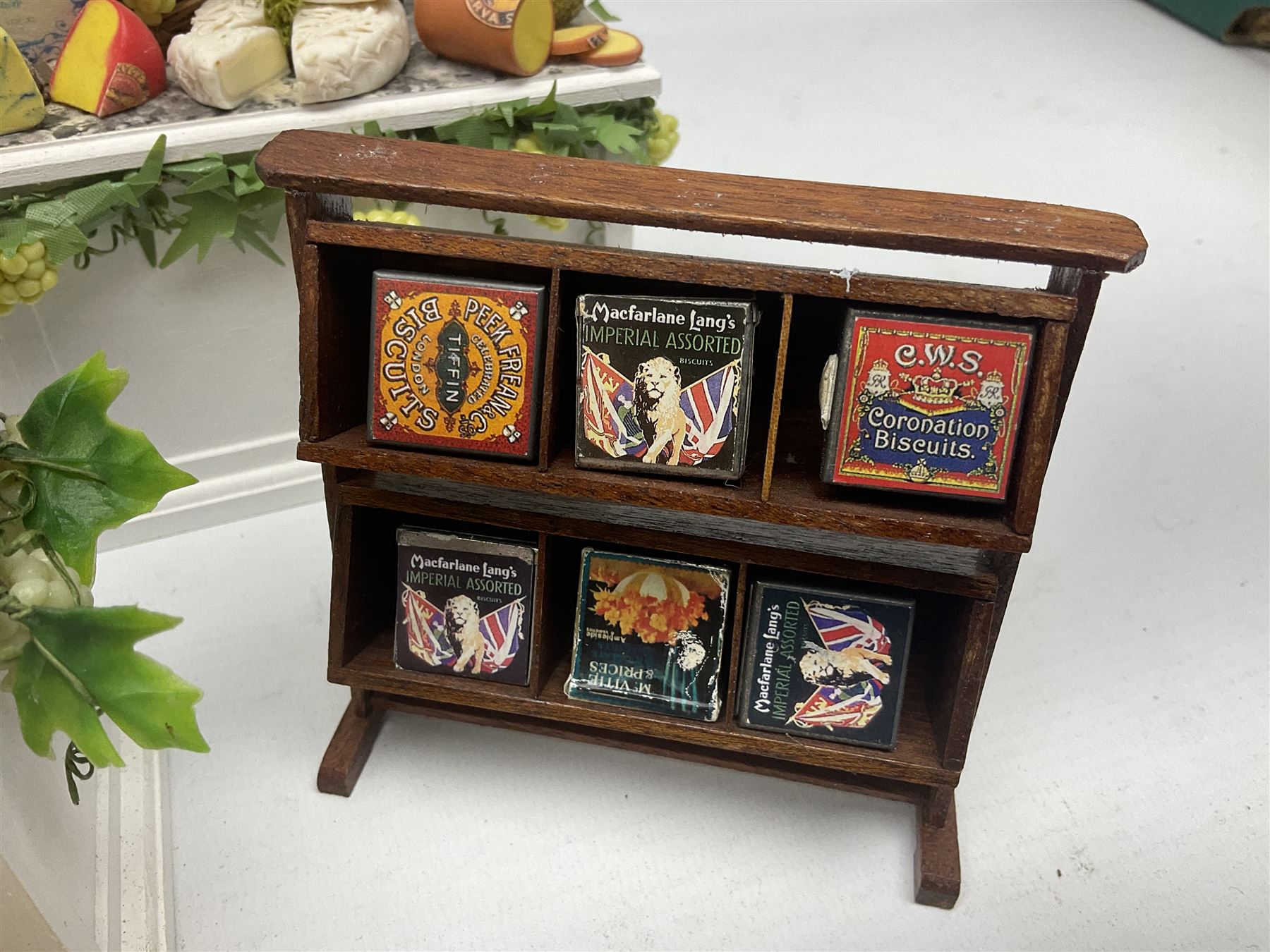 Collection of miniature dolls house grocery shop furniture and produce, comprising glazed corner counter with brass style cash register, shelved preserves unit, pies, cheese board and trays of meats, pair of white cheese counters, biscuit display with six tins, glazed preserves cabinet, pair of shop chairs and four further cheese boards complete with grapes and label flags