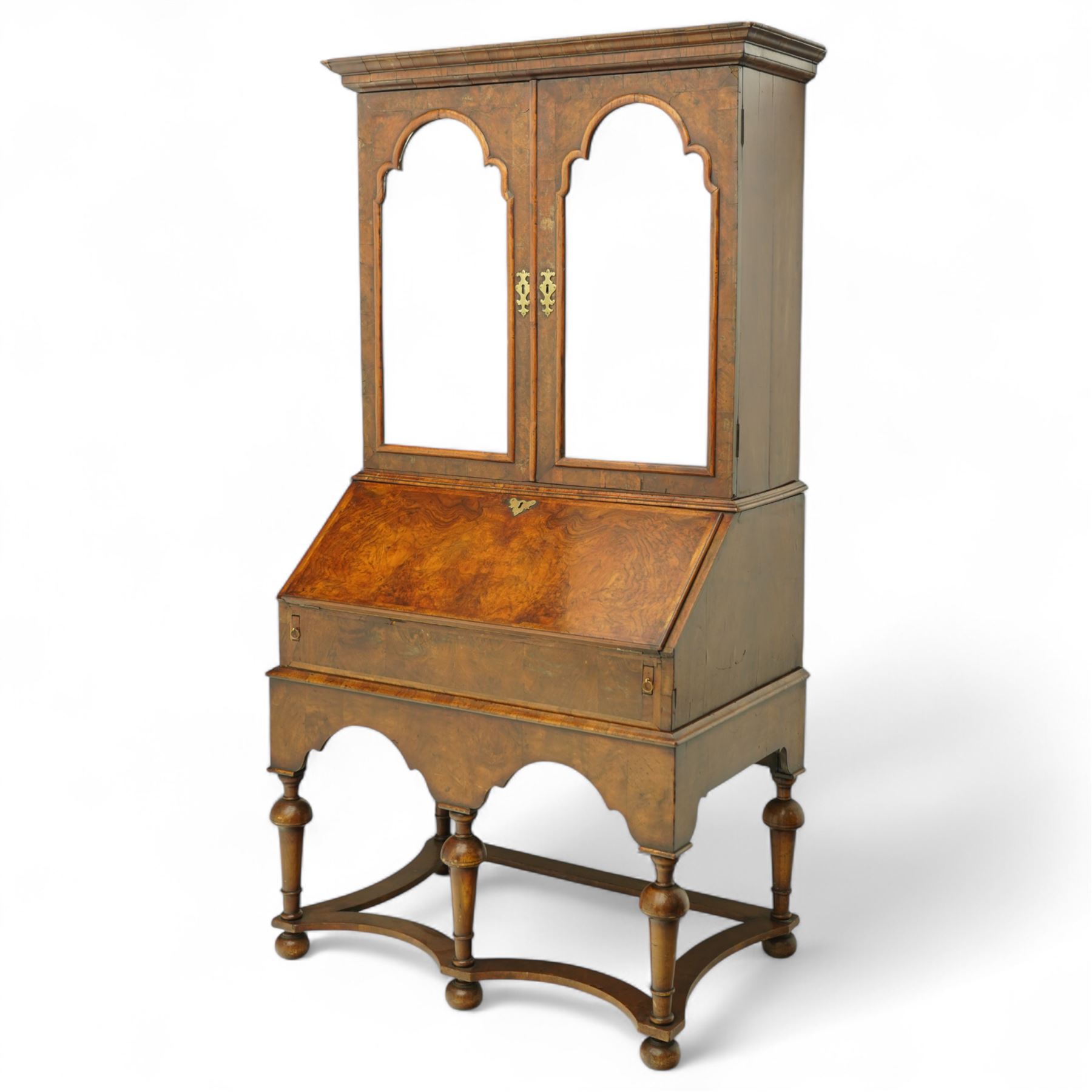 Queen Anne burr walnut chest on stand, moulded projecting cornice over arched mirror glazed doors, above a sloping fall front enclosing fitted interior with pigeonholes and correspondence drawers, fitted with foliate brass escutcheons, shaped apron over tapered baluster turned supports united by curved stretchers, early 18th century with later restorations 
