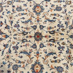 Large Central Persian Kashan hand-knotted carpet, ivory field with an all-over scrolling design of vines, palmettes, and floral sprays, enclosed by a broad camel-ground palmette and vine border, framed by multiple guard stripes in a palette of ivory, beige, blue and polychromed tones, with cotton fringe ends 