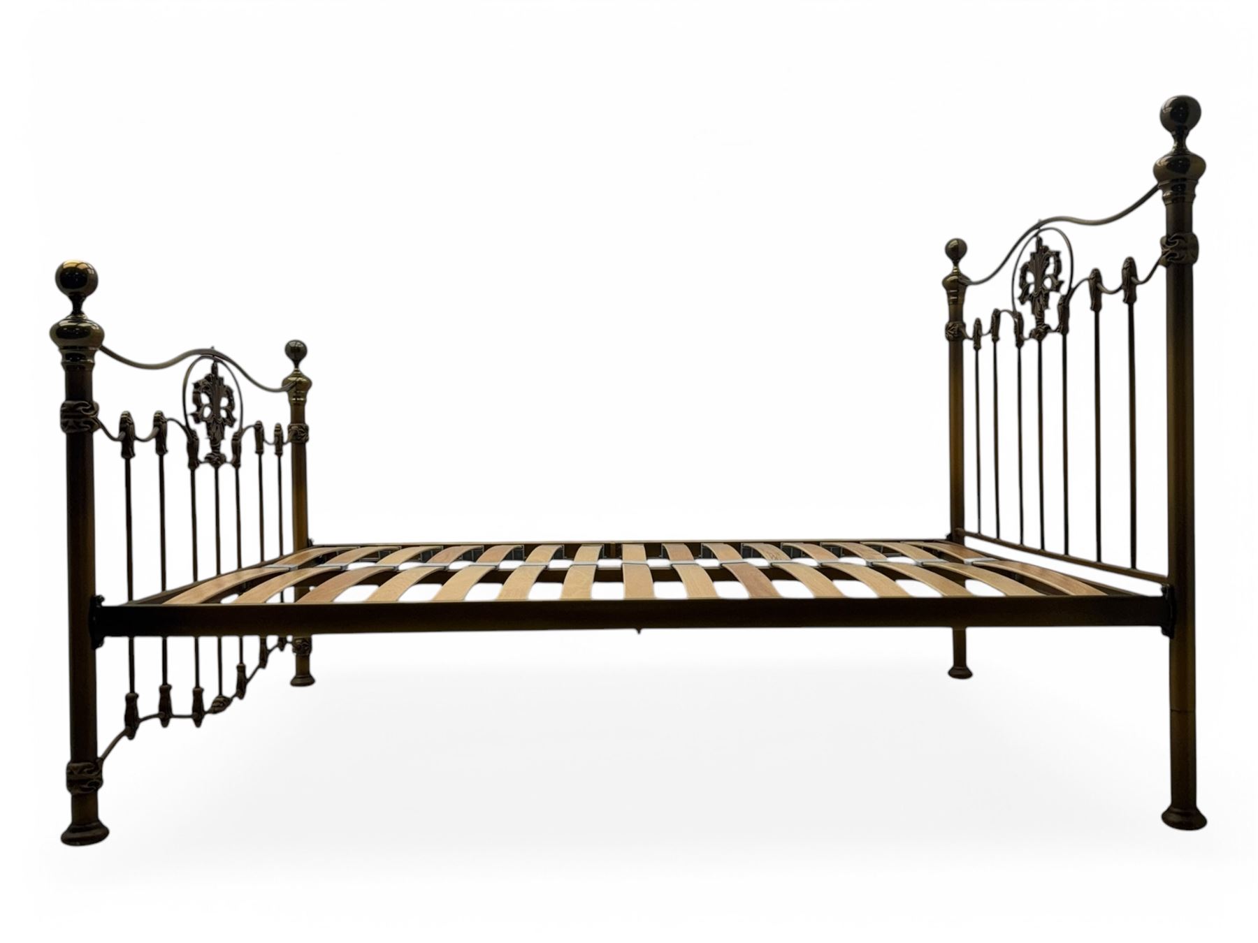 Victorian design king size brass bed frame, featuring an arched headboard and footboard with intricate cast floral and scrollwork centre motifs, flanked by cylindrical supports with ball finials and decorative collars, raised on rounded feet, with a slatted wooden base