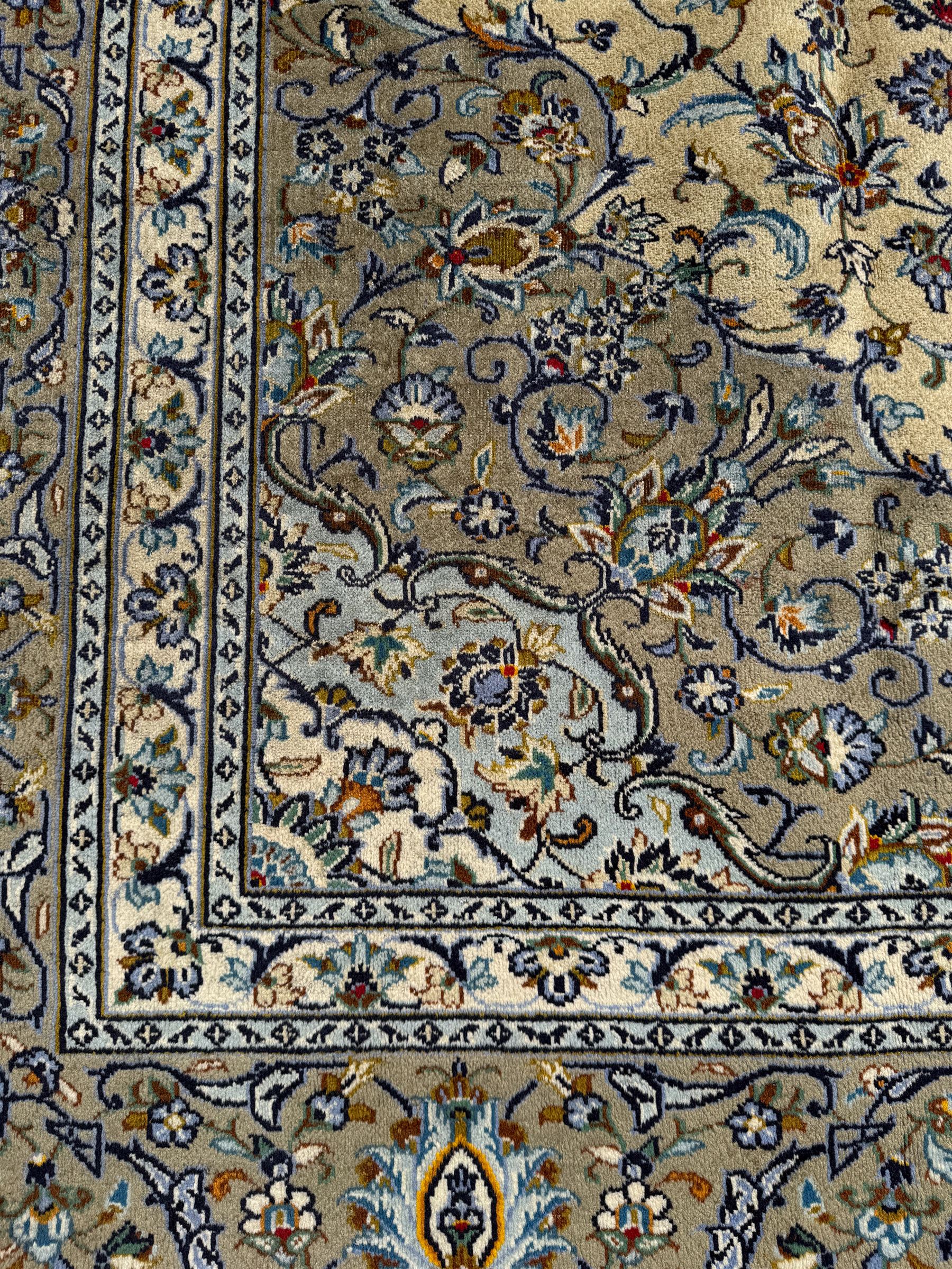 Central Persian Kashan pale khaki and ivory ground carpet, shaped central medallion enclosing small stylised motifs, the field decorated profusely with trailing foliate branches and plant motifs, guarded border with repeating floral design