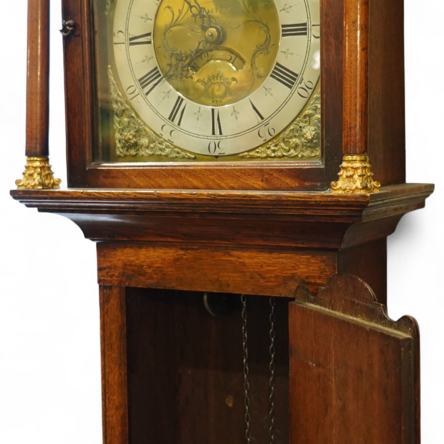 Gilbert Kidd of Malton-oak cased mid-18th century 30hr longcase clock, with a caddy top and three gesso finials, square hood door flanked by turned pilasters with Corinthian capitals, trunk with a long wavy topped door on a square plinth with an shaped skirt, brass dial with an engraved dial centre, date aperture and steal hands, silvered chapter ring with Roman numerals and five minute Arabic's, dial pinned to a chain driven countwheel striking movement. With a brass cased weight and pendulum. 
