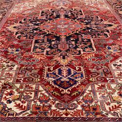 North West Persian Heriz hand-knotted carpet, madder red field with large central stepped medallion with angular floral and geometric designs, surrounded by stylized palmettes, leaves and tribal motifs with indigo corner spandrels, enclosed by a broad navy border of geometric floral devices between multiple guard stripes, with cotton fringe ends 