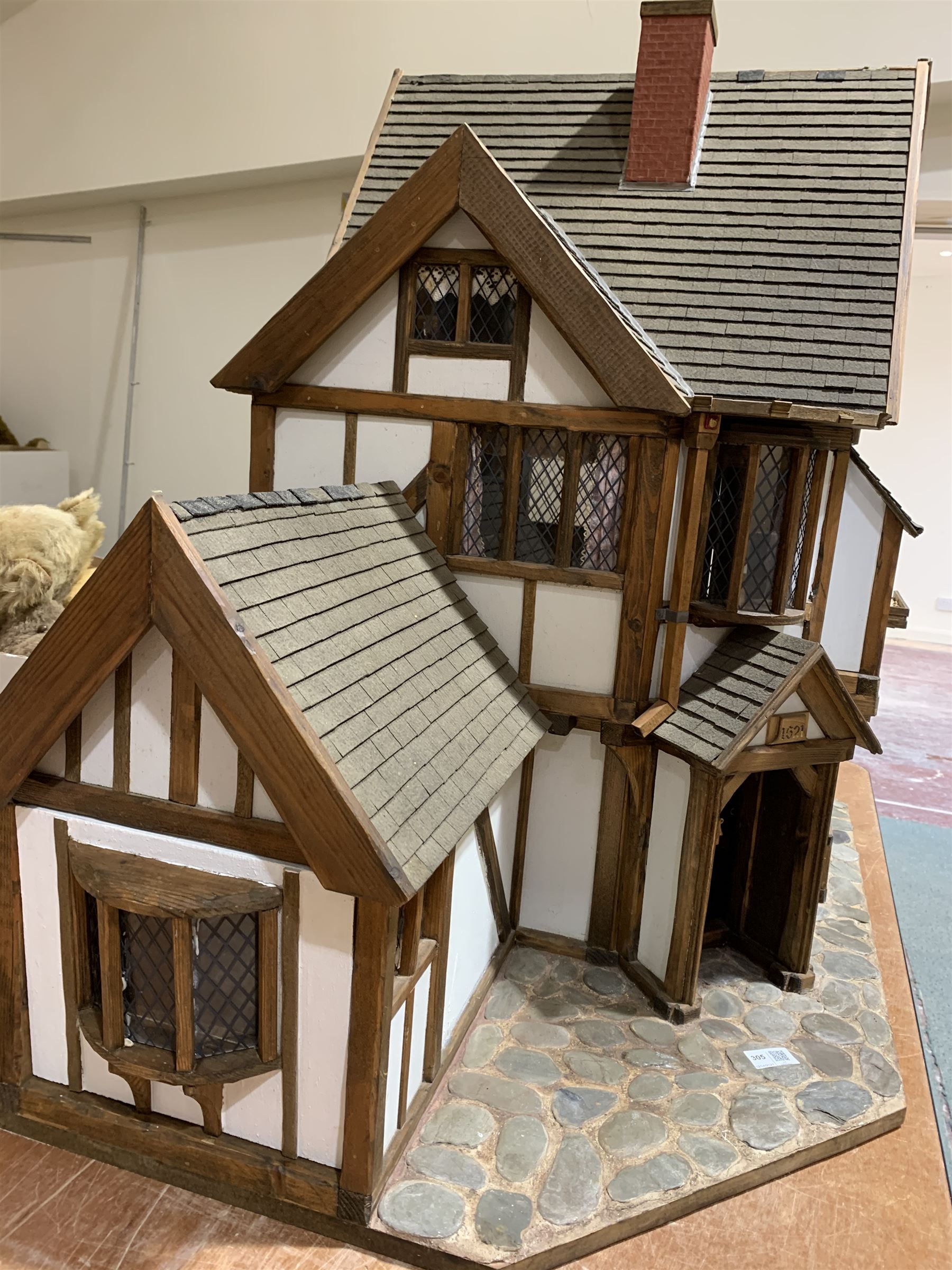 Gerry Welch for Manorcraft Tudor style wooden doll's house, of three-storey multi gabled form with mock beam and plaster walls and simulated leaded glass windows under a felt tiled roof, boarded floors and stairs to each room, curtains to most windows, front entrance door, rear door and single storey extension for kitchen. Furniture includes fitted floral bathroom suite to top floor, well stocked Welsh dresser with copperware etc, tapestry style wall hangings, knole settee, tin bath, metal range, fire-surround, wooden three-piece suite etc; on shaped cobbled base W91cm D61cm H91cm