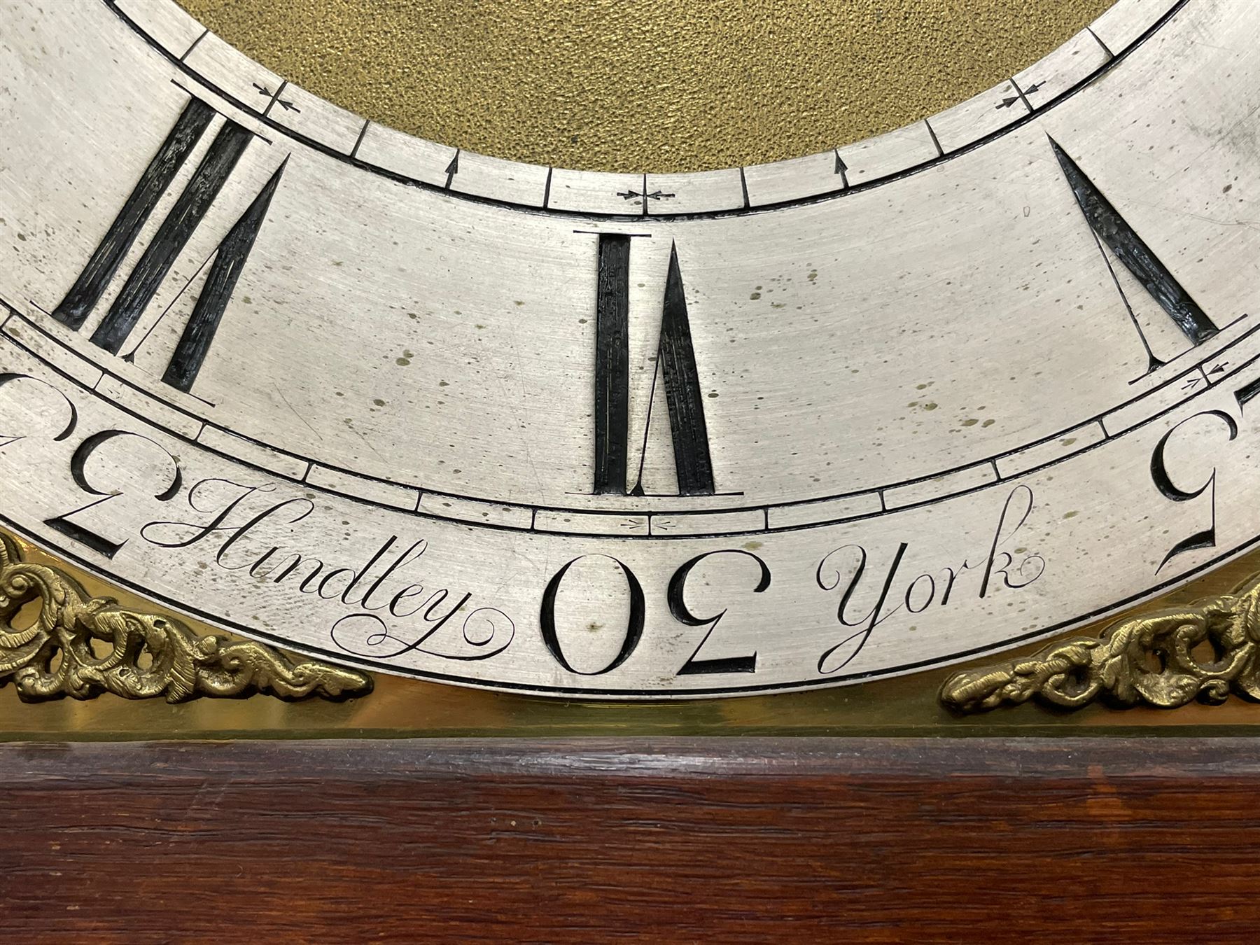 Henry Hindley of York – mid-18th century oak cased 8-day longcase clock,
flat topped caddy pediment with gesso finials, rectangular hood door flanked by wooden turned pilasters with capitals, canted trunk with a full-length break arch topped door on a short plinth with applied skirting and bracket feet, brass dial with a silvered chapter ring, Roman numerals and five-minute Arabic’s, minute and inner quarter hour tracks, matted dial centre with subsidiary seconds dial, blued steel hands and female head pattern spandrels, silvered calendar dial to the break arch with dolphin spandrels, four pillar rack striking movement, striking the hours on a bell. With weights and pendulum. 
H250 W48 D27
Henry Hindley originally from Wigan (Lancs) is recorded as working in York from Petergate 1731-41 then Stonegate 1741-1771. A famous and highly innovative maker, often referred to as the “Tompion of the North.” 

