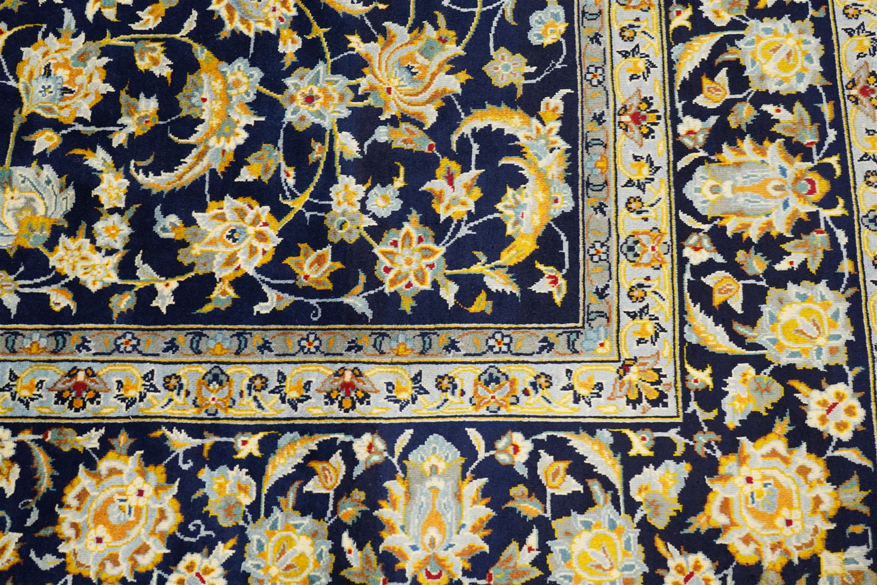 Central Persian Kashan hand-knotted carpet, deep indigo field with a densely all-over arrangement of scrolling floral vines, palmettes, and flowering sprays, in soft ivory, pale blue, and warm gold tones, principal ivory border with a continuous meandering vine of stylized palmettes, and floral motifs, flanked by multiple narrow guard borders, with cotton fringe ends 