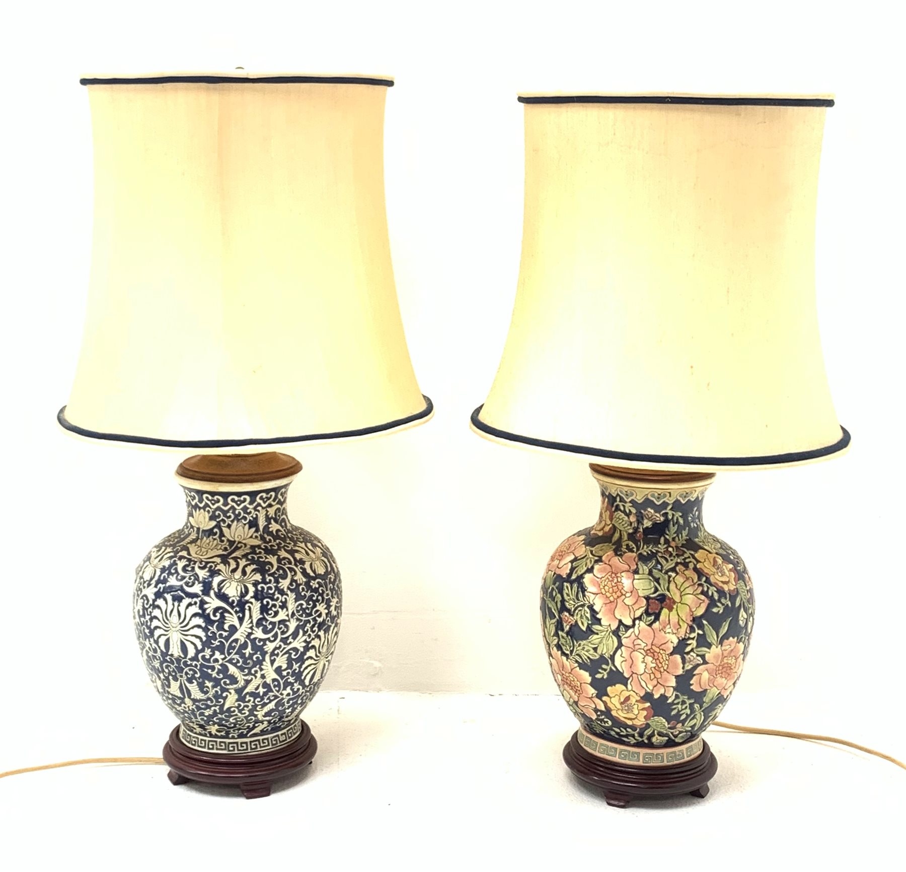 Two Oriental table lamps, each of bulbous form, the first example decorated with pink blossoming flowers upon a dark blue ground, above a fret pattern band, upon a wooden circular stepped base, the second example with white foliate decoration upon a dark blue ground, also with fret pattern band, and upon a wooden circular stepped base, each with fabric shade, including shade each approximately 74cm. 

