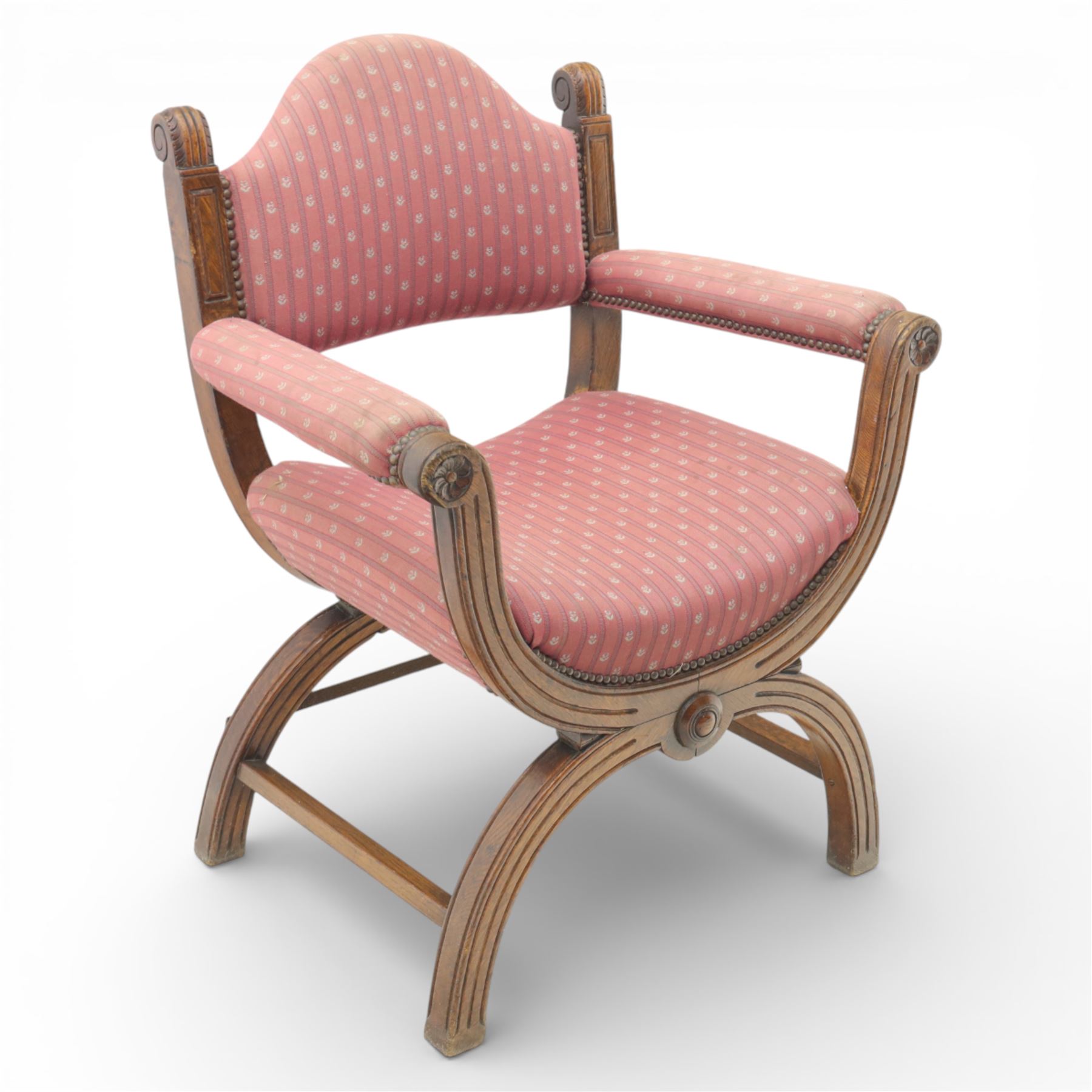Late Victorian oak x-frame armchair, upholstered padded back and seat covered in pink patterned fabric with close studded borders, shaped downswept arms with scroll terminals, raised on curved x-frame supports with moulded scroll feet