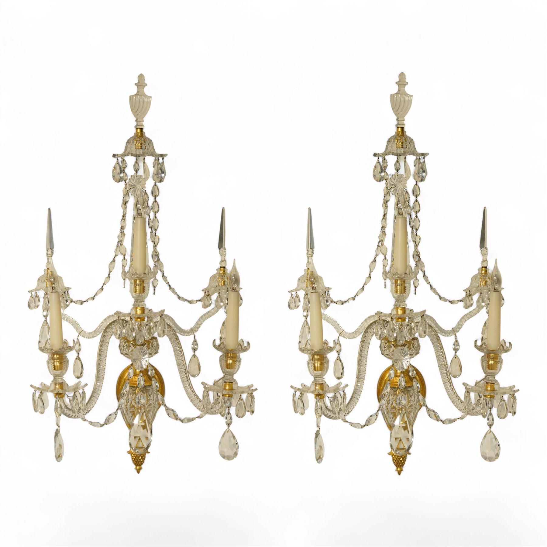 Pair of George III ormolu and cut-glass wall-lights, each with a tall central urn-shaped finial above a series of faceted glass spires and scrolled branches, the ormolu-mounted nozzles and drip pans hung with festoons of cut-glass drops and pear-shaped pendants, the lower section centred by a pineapple finial, wired for electricity