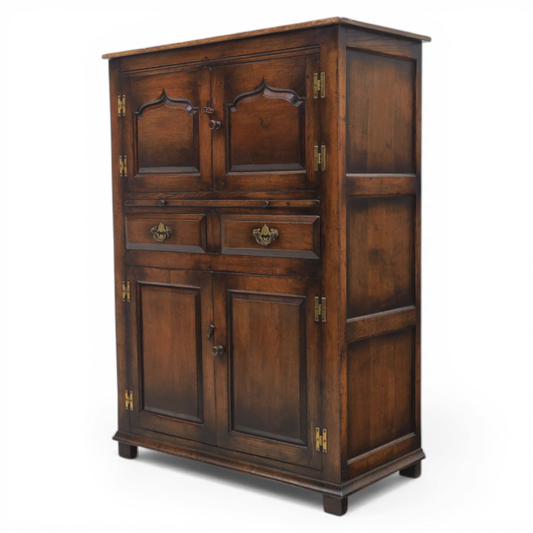  Late 20th century oak four door cupboard in the manner of Titchmarsh and Goodwin, rectangular plank top above two pairs of fielded panel doors with shaped raised panels, central arrangement of a brushing slide above two short drawers with brass swan neck handles and pierced backplates, enclosed by further pair of panelled cupboard doors, fitted with brass butt hinges and turned wooden knobs, raised on plinth base