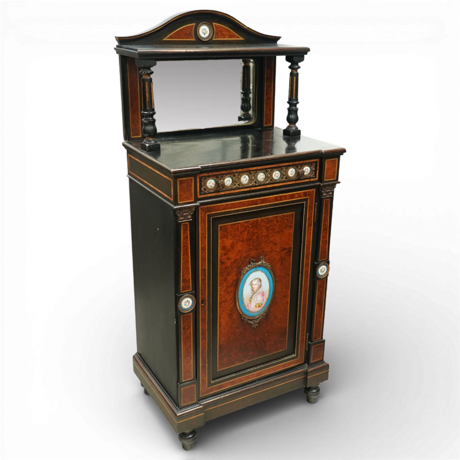 Victorian Aesthetic Movement amboyna and ebonised cabinet, the arched raised back inlaid with amboyna and floral porcelain plaque, over a raised shelf with fluted and turned supports, and a bevelled mirror plate with brass beading beneath, the inverted-breakfront top over an ornate frieze drawer with guilloche style gilt-metal mounts and porcelain plaques, the panelled door with a central porcelain plaque depicting a lady in a fuchsia gown within concentric ebonised and amboyna inlays, flanked by architectural style uprights with Corinthian capitals, raised on turned feet