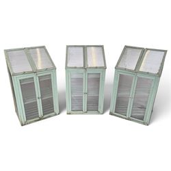 Three green-painted timber cold-frame greenhouses or growhouses, each with twin hinged sloping polycarbonate lids above a pair of panelled doors enclosing slatted shelves