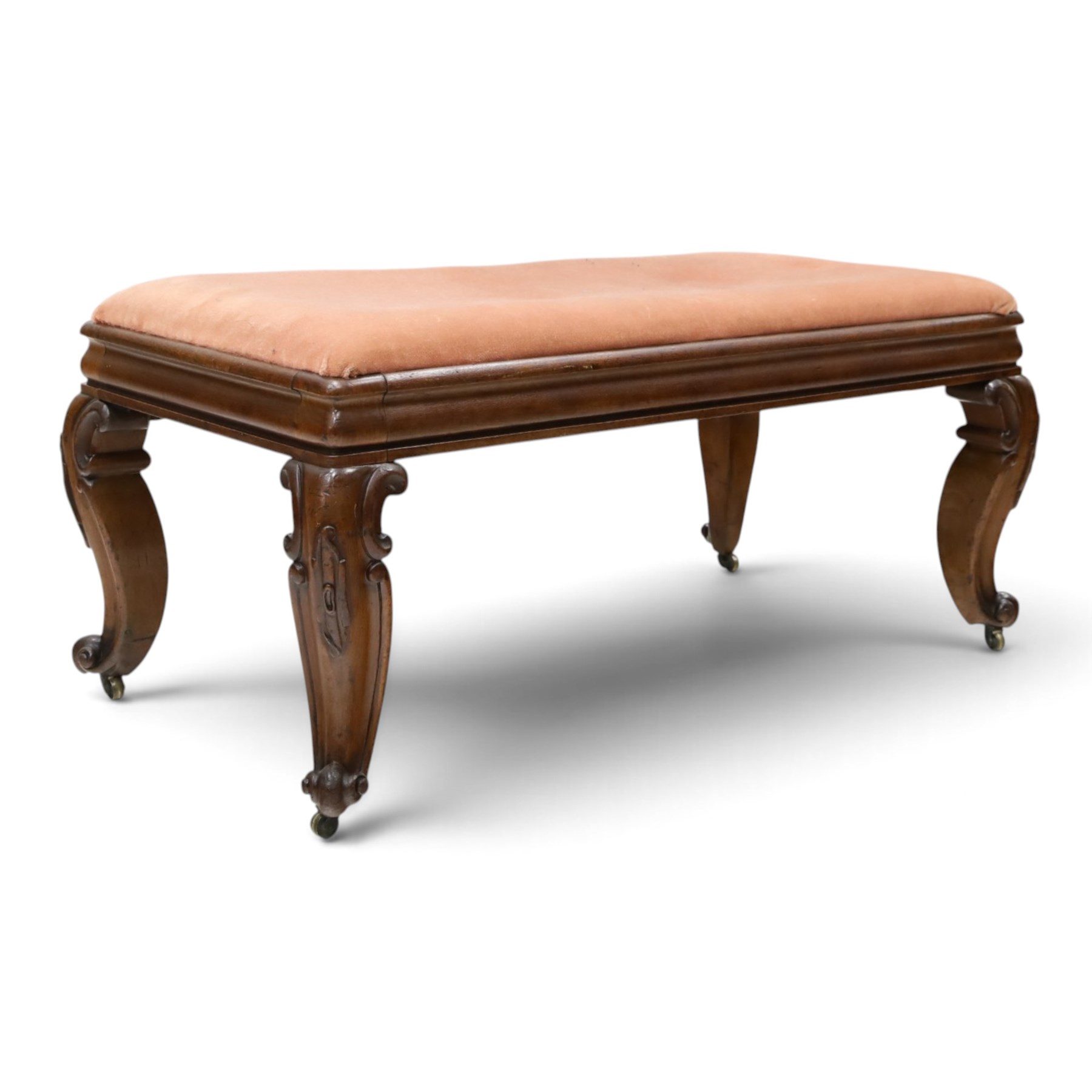 Victorian carved mahogany fender stool of generous proportions, the wide rectangular seat upholstered in rose fabric, shaped frieze rail over C-scroll and cartouche carved cabriole supports with scroll feet and brass castors