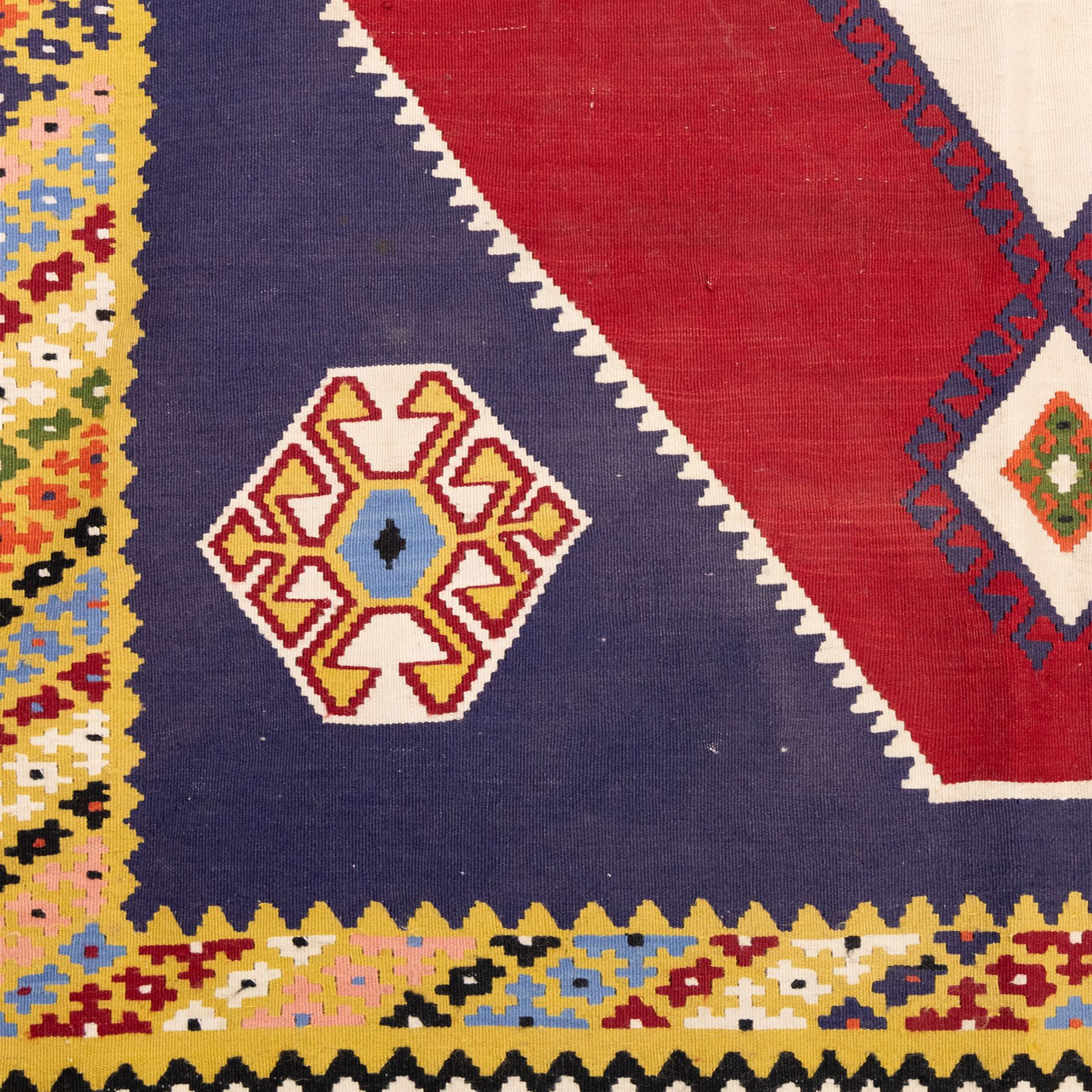 South West Persian Qashqai kilim, woven in flatweave with tones of indigo blue, madder red, yellow, turquoise, and green, with a bold central diamond medallion on an ivory ground, enclosing smaller lozenge motifs and flanked by deep madder red panels, field framed by a broad indigo border with polychrome geometric devices and serrated motifs 