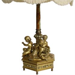 Second Empire style gilt metal table lamp, the hexagonal plinth base with applied foliate decoration, upon six turned feet, supporting three seated allegorical putti, the first modelled with a tablet and stylus representing literature, the second with hammer representing sculpture, and the third with Corinthian column capital representing Architecture, each intersected by a stylised scroll with central flower head panel, mounted upon an onyx and gilt metal Corinthian column with wrythen flower moulded and part fluted base, supporting a cream octagonal shade, overall H86cm