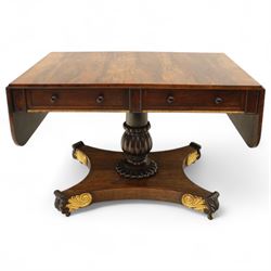 Regency rosewood sofa table, rectangular top with variegated rosewood veneers, hinged D-shaped leaves, over a set of dummy drawers to the back and full-size drawers to the front, with turned knob handles and gilt beaded moulding on frieze above elegant turned, tapering, and fluted, pedestal with a well-carved collar, supported on a large, tripartite concave platform base terminating in richly carved lion paw feet with gilded acanthus leaf carving, on concealed brass castors, circa 1815, length extended 161cm 
