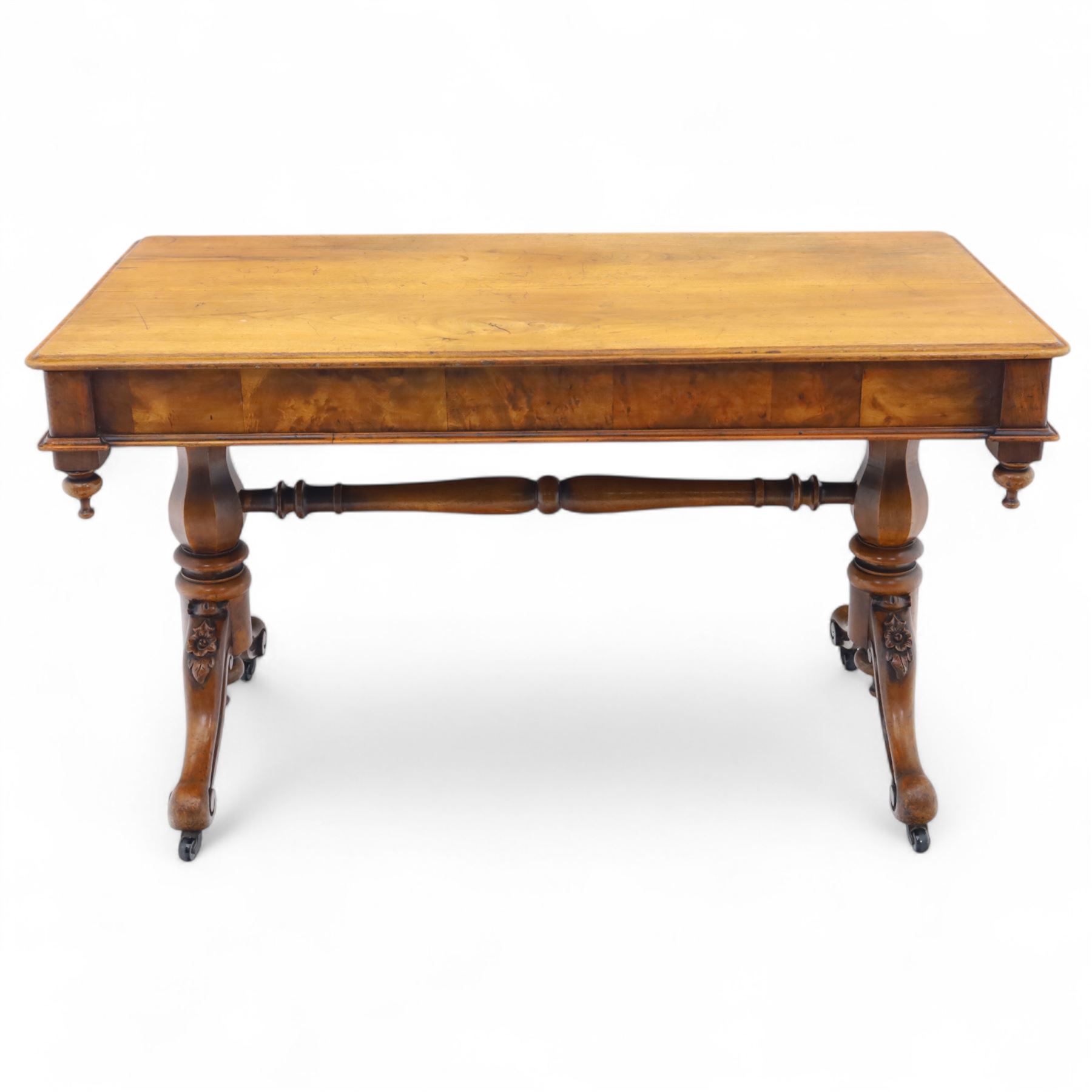 Mid Victorian walnut library table, plain top of rectangular form with moulded edge above frieze fitted with opposing drawers, table supported on a pair of turned and hexagonal carved baluster end standards, united by a turned stretcher terminating in scrolling sabre legs carved with acanthus and floral details, resting on ceramic castors, circa 1860