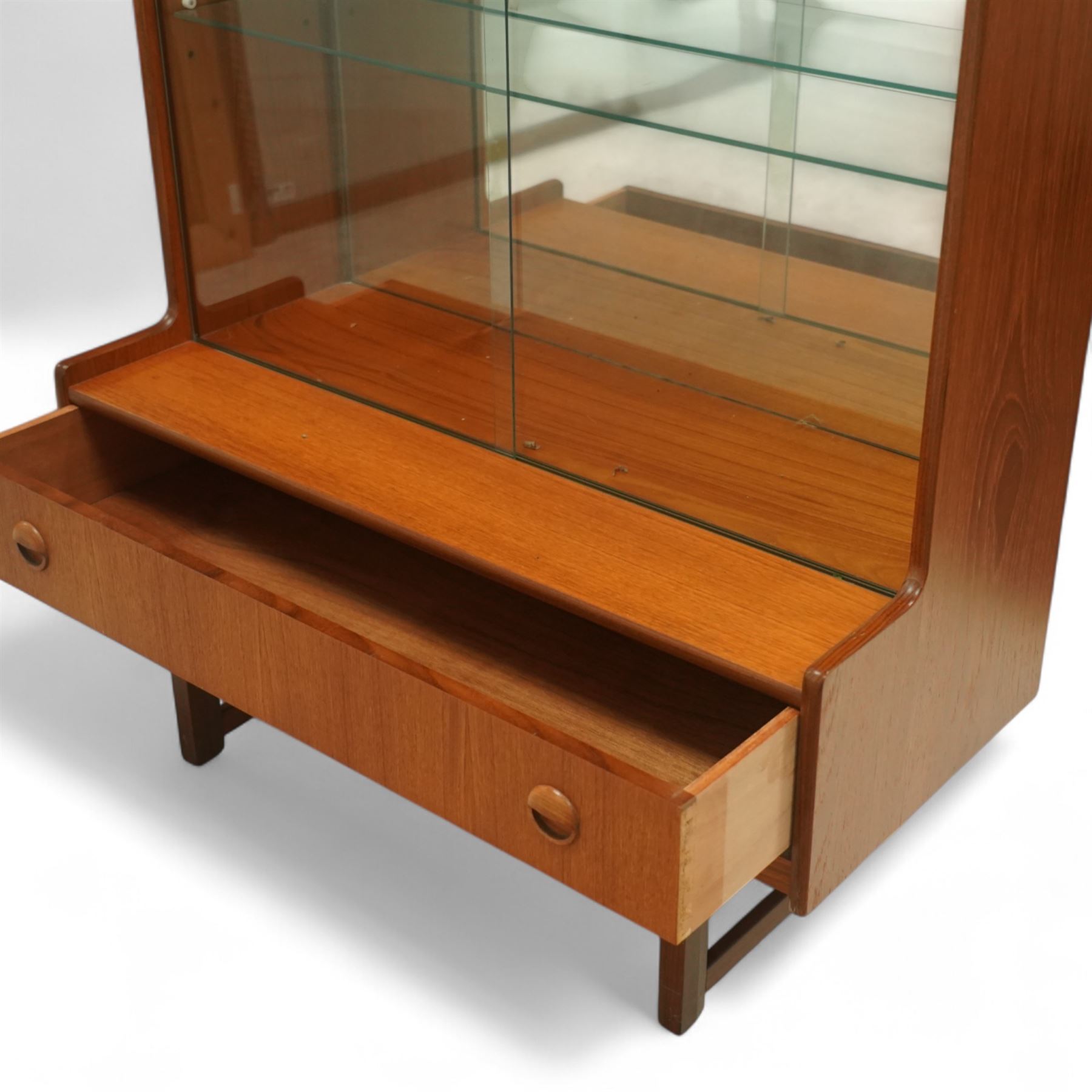 Turnidge of London mid-century teak display cabinet, rectangular top with a pair of glass sliding doors enclosing two adjustable shelves with mirrored back, projecting lower section incorporating one full-width drawer with recessed circular handles, raised on square-section legs with lateral stretchers  