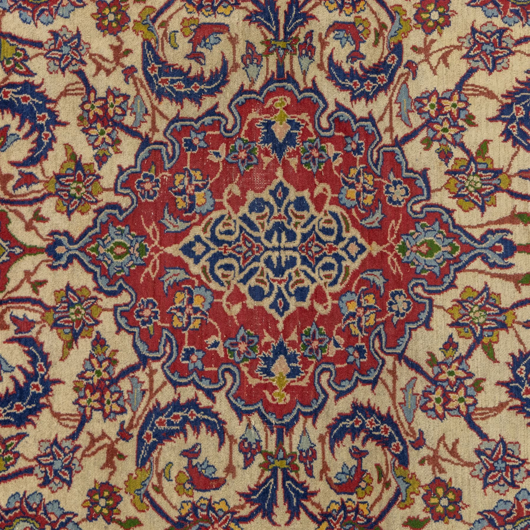 Kashan design wool rug, with red floral medallion on a cream field, decorated with scrolling foliate motifs in blue, red and green tones, enclosed by red and blue floral borders with fringed ends
