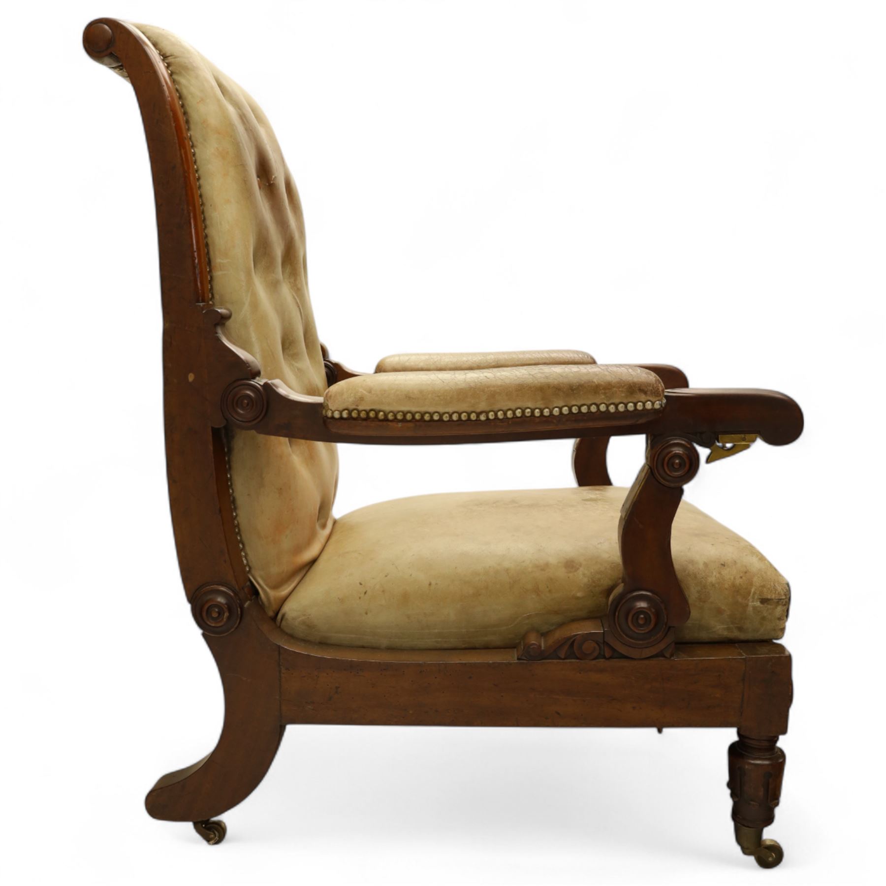 William IV mahogany gentleman's library reclining armchair, leather button upholstered and brass-studded back and seat, above leather-covered arms terminating with carved roundels and foliate scroll-carved ends, underside with brass mechanical trigger reclining lever, the apron with retractable foot rest, supported on lappet carved and turned legs with brass castors, circa 1835