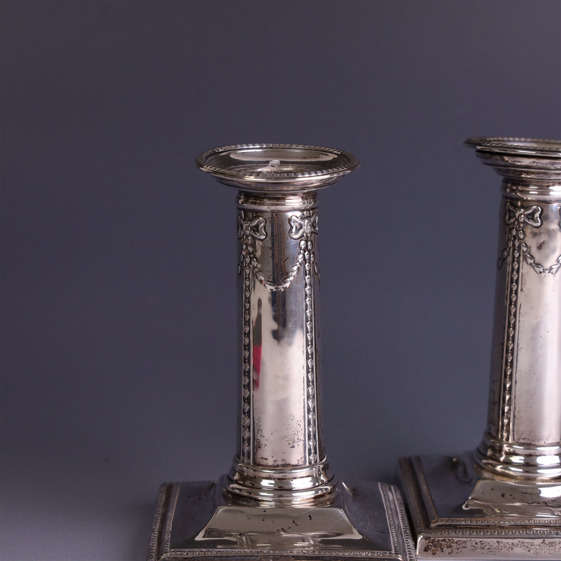 Pair of late Victorian silver mounted candlesticks, the stems embossed with classical ribbon and swag design, upon weighted stepped square bases, hallmarked London 1899, maker's mark worn and indistinct, H12cm