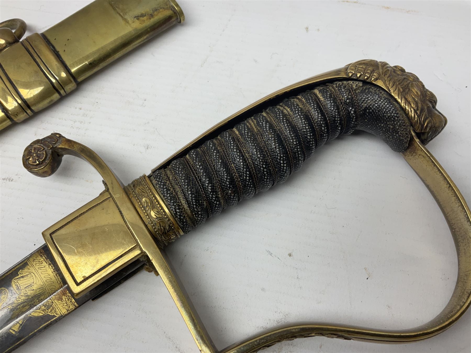 German Saxon 1st Empire Light Cavalry officer's sword c1820s, the 79cm clipped curving blade engraved with the crest of Hanover and various battle trophies with traces of blueing and gilding, brass hilt with knucklebow, foliate square langets, ornate downswept quillon, wire-bound fish skin grip with lion's head pommel; in brass scabbard with two suspension rings L95cm overall