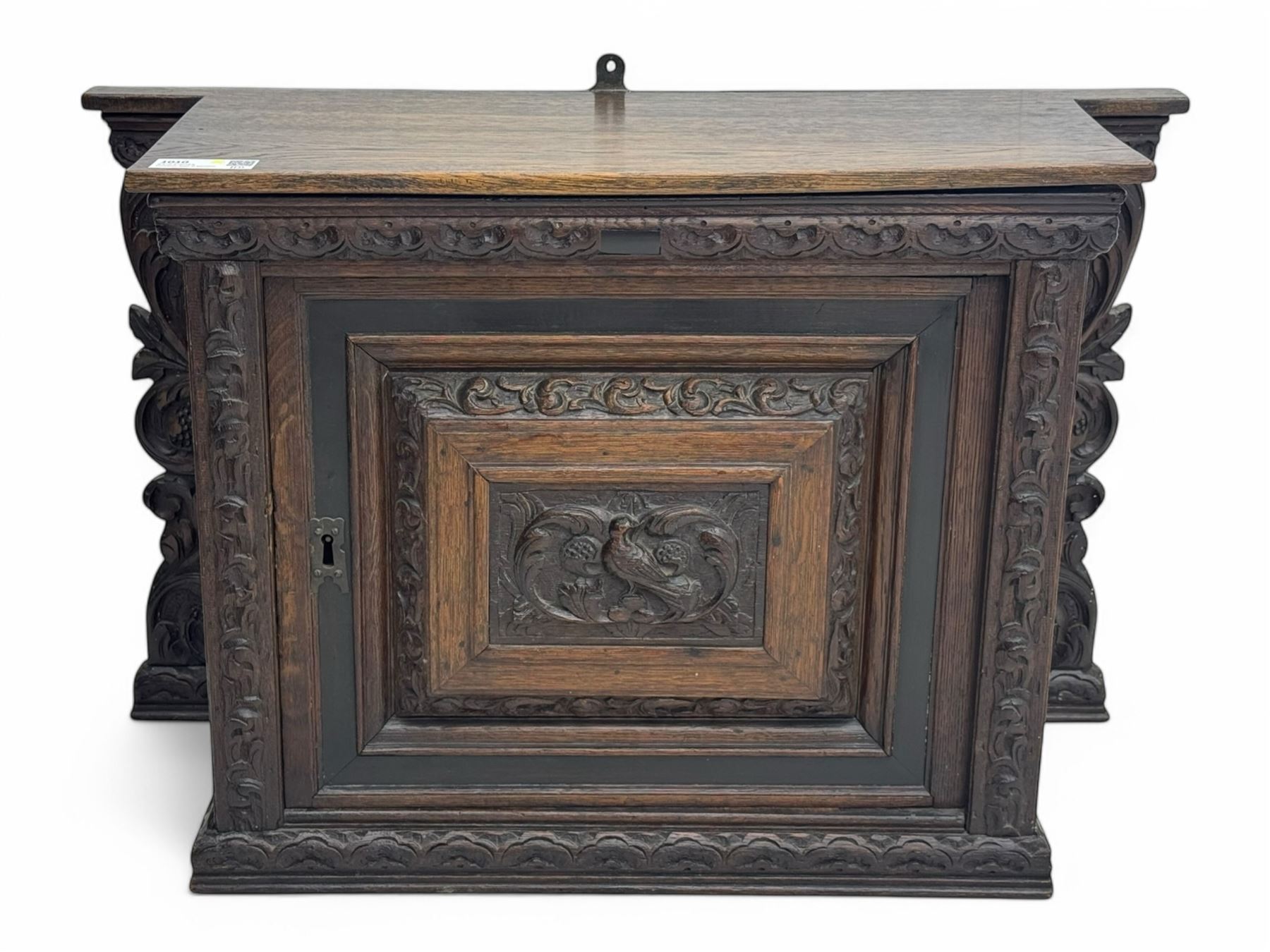 18th century and later carved oak cupboard, enclosed by panelled door carved with central bird motif and scrolled leafage with an outer ebony band, the frieze and uprights carved with repeating lunette and floral designs, flanked by scrolled foliate carved upright brackets