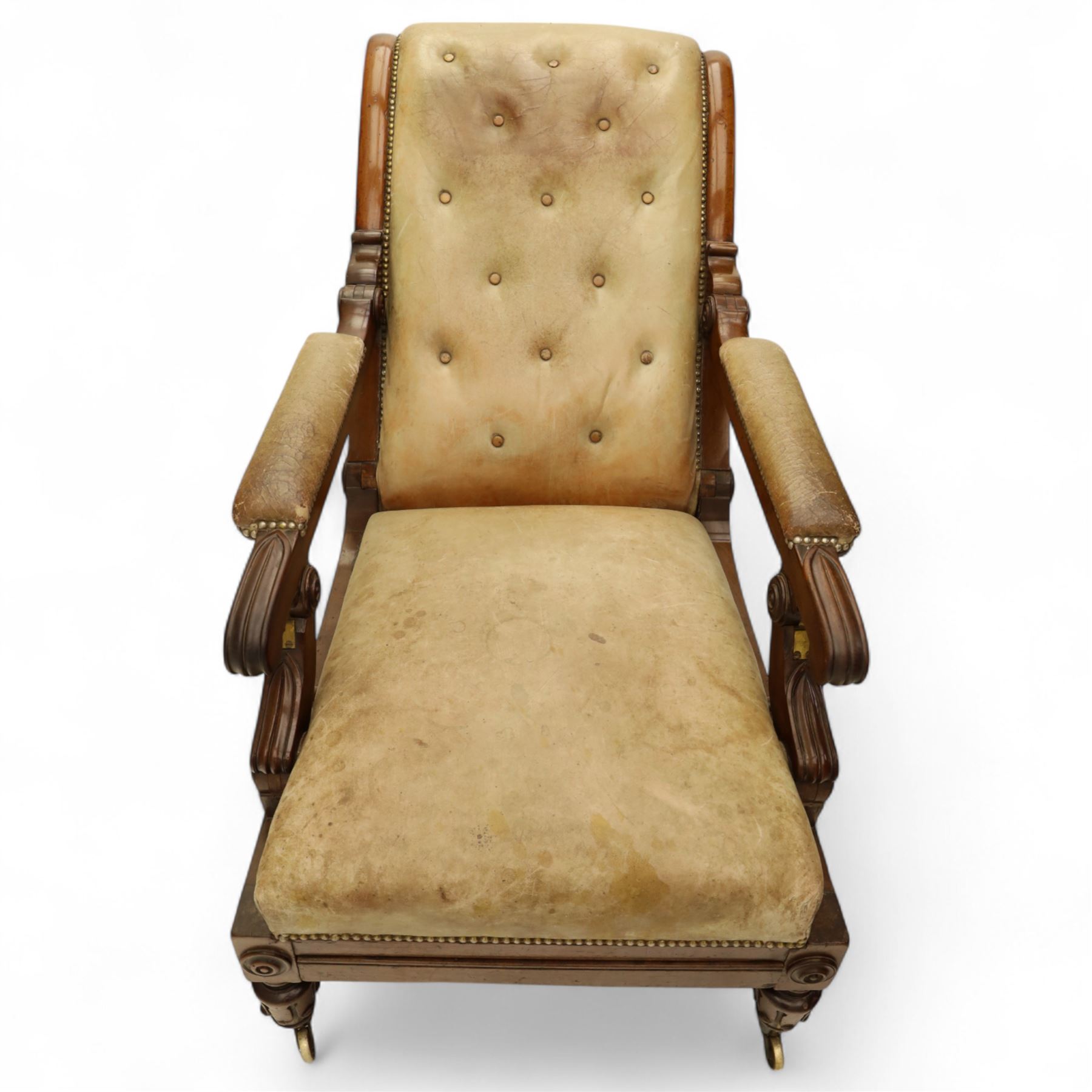 William IV mahogany gentleman's library reclining armchair, leather button upholstered and brass-studded back and seat, above leather-covered arms terminating with carved roundels and foliate scroll-carved ends, underside with brass mechanical trigger reclining lever, the apron with retractable foot rest, supported on lappet carved and turned legs with brass castors, circa 1835