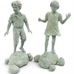After Eneri Prosperi, bronze garden child statues, depicting children balancing on rock-form bases, the boy in shirt and shorts, the girl in dress with raised arms, surrounded by loose rounded pebble elements