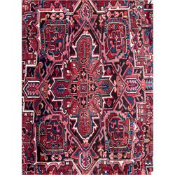 Northwestern Persian Heriz hand-knotted carpet, red madder field with a bold stepped medallion in indigo, ivory, and terracotta, flanked by angular floral motifs and stylized leaves, the spandrels set within a dark indigo border of rosettes and angular vine motifs between narrow guard stripes, 340cm x 245cm 