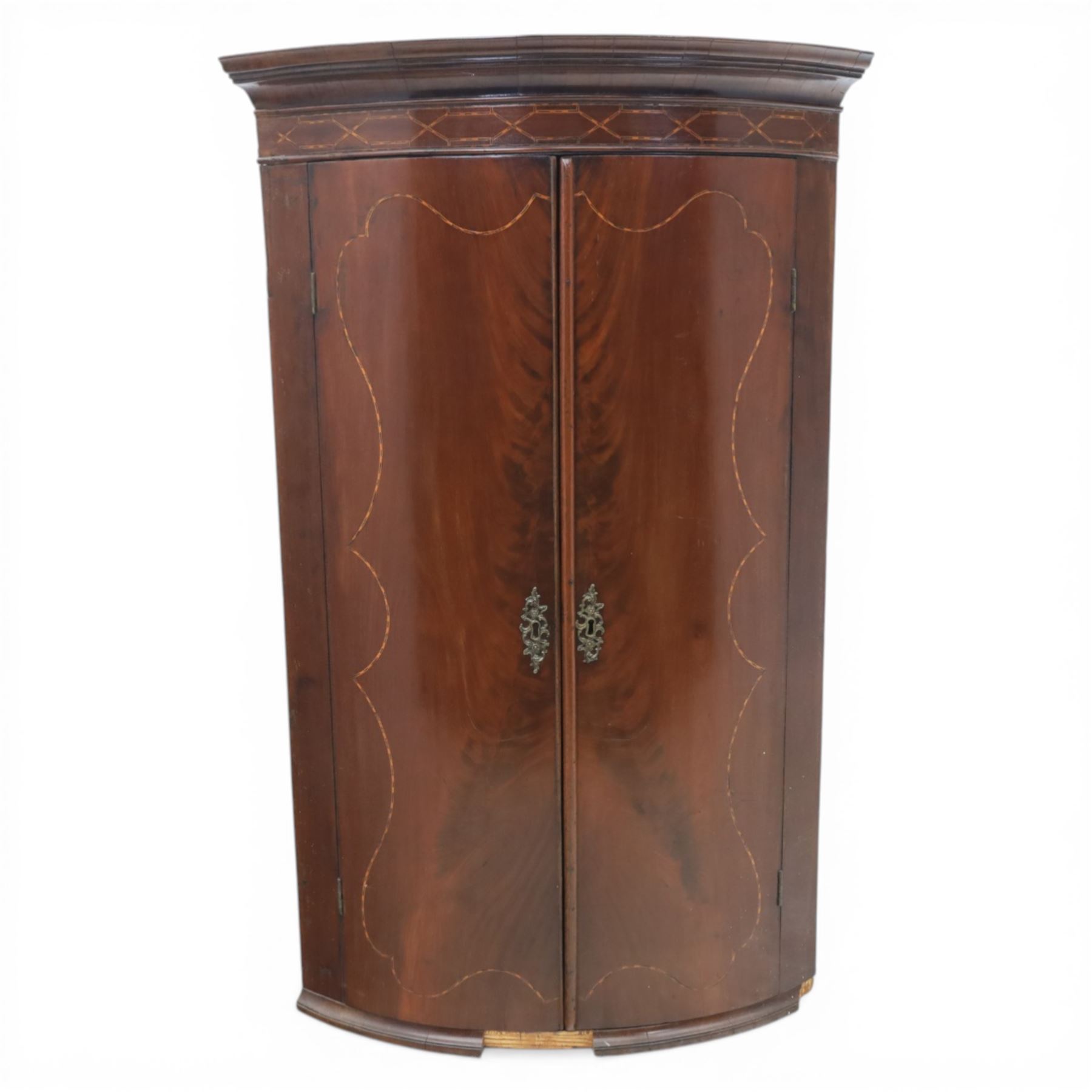 George III mahogany bow-front corner cupboard, projecting moulded cornice over satinwood inlaid frieze with geometric crossbanding, enclosed by twin shaped panel doors with line inlay and ornate brass escutcheon plates, painted interior fitted with shaped shelves, on moulded plinth base