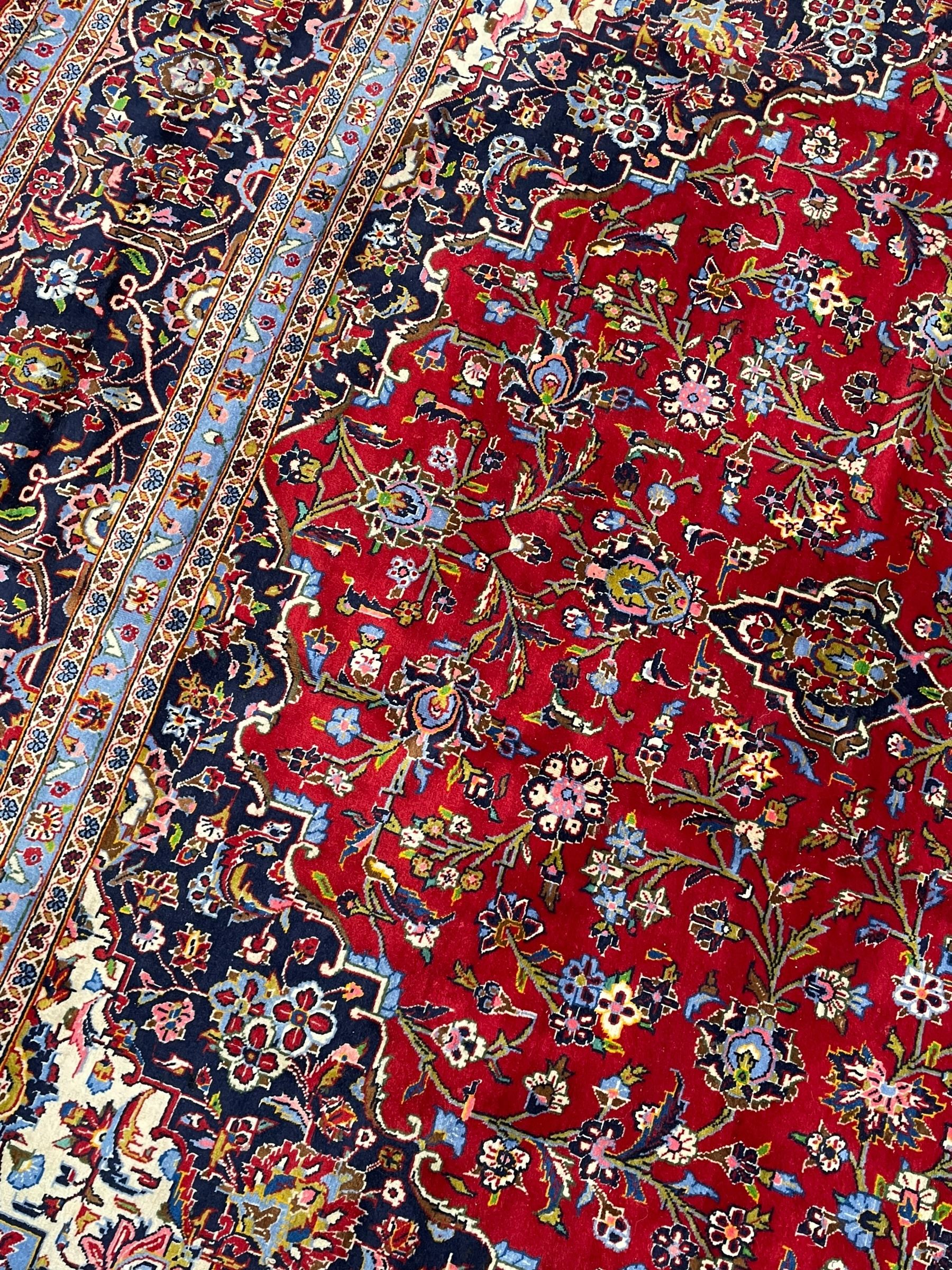Persian Kashan crimson ground carpet, floral pattern indigo ground central medallion and matching spandrels, the field decorated densely with scrolling foliate patterns and floral motifs, the main border decorated with scrolling branches and palmettes, within floral pattern guard stripes