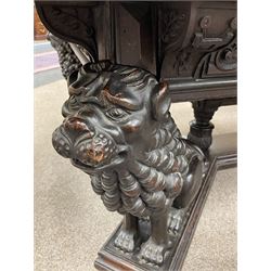 Large 19th century carved oak library table, the rectangular top with lunette carved edge over cartouche panelled and carved  scroll frieze, the central tapering rectangular support relief carved with masks and garlands, on four carved seated lion supports at each corner, on flat angular stretcher with recessed castors stamped ‘Bartholomew & Fletcher, London’