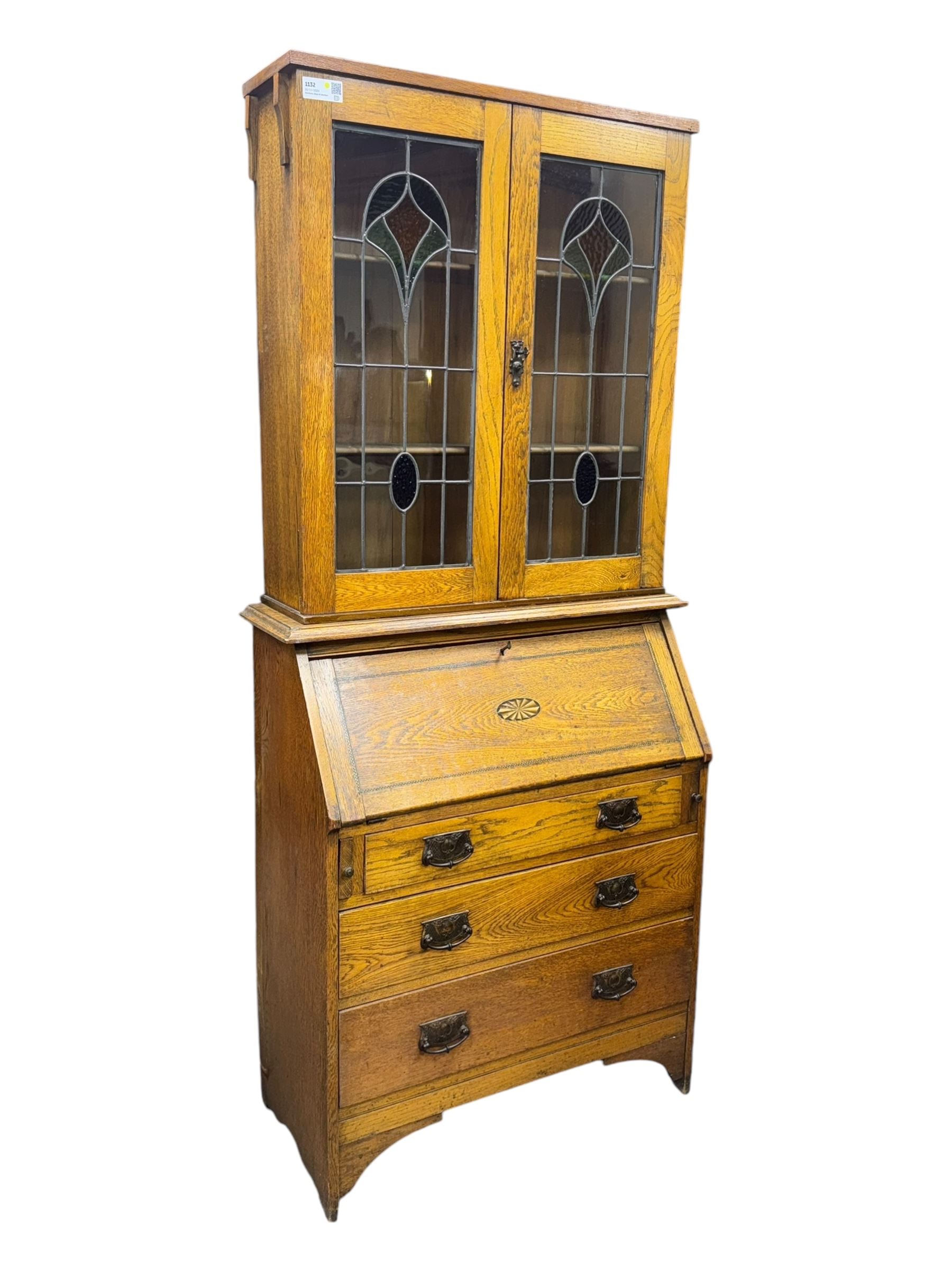 Early 20th century Arts and Crafts oak bureau bookcase, twin leaded and glazed stained glass doors enclosing two shelves, over bureau section with fall-front inlaid with sunburst motif, fitted interior of pigeonholes and shelves, on bracket feet