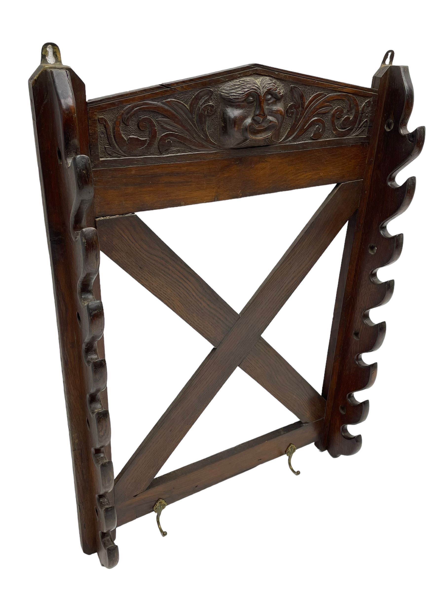 Victorian oak wall hanging whip or stick rack, sloped arch pediment carved with central grotesque mask with extended scrolling foliage, the upright supports cut with rests, x-framed back, two lower hooks