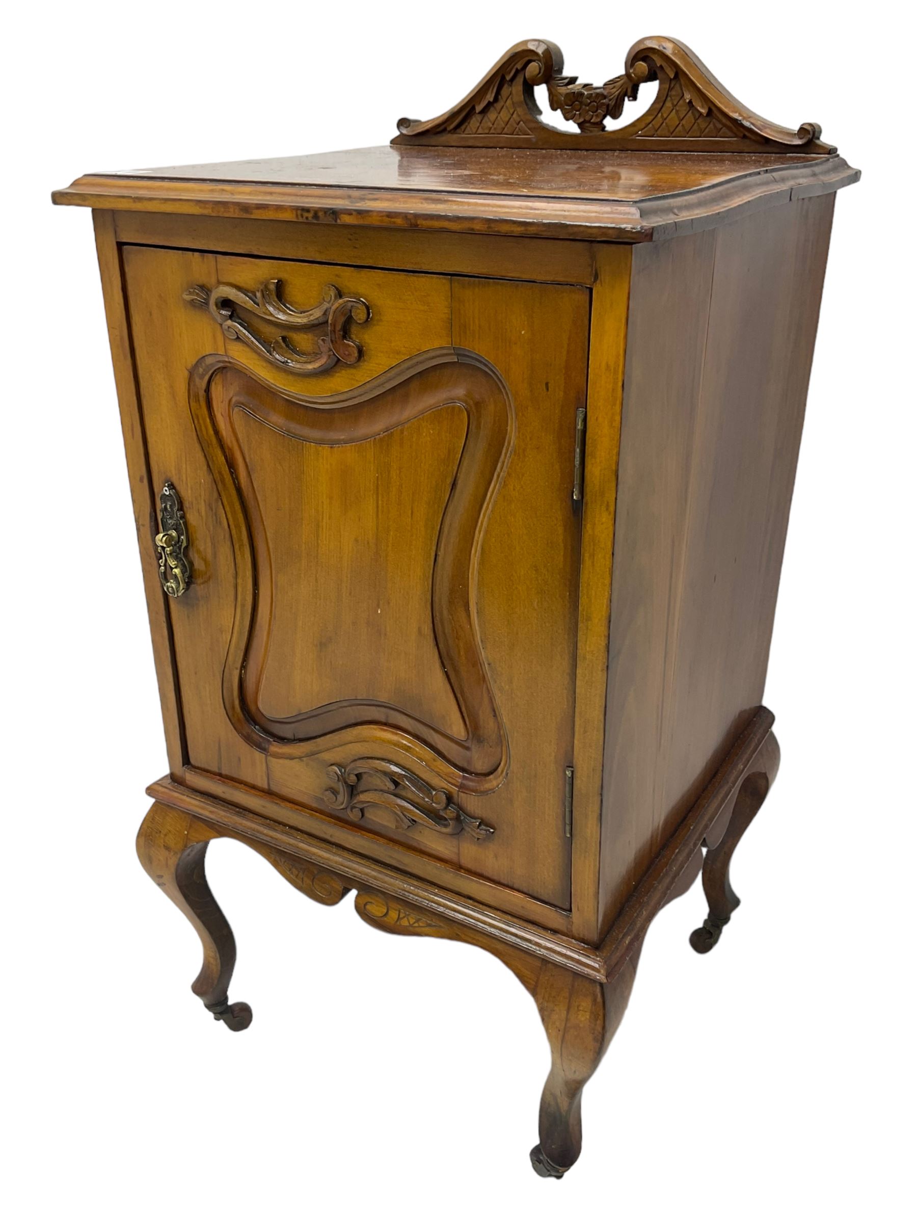 Edwardian Art Nouveau walnut bedside cabinet, raised broken swan neck back with carved flower heads, single fielded panelled door enclosing single shelf, raised on cabriole supports with ceramic castors