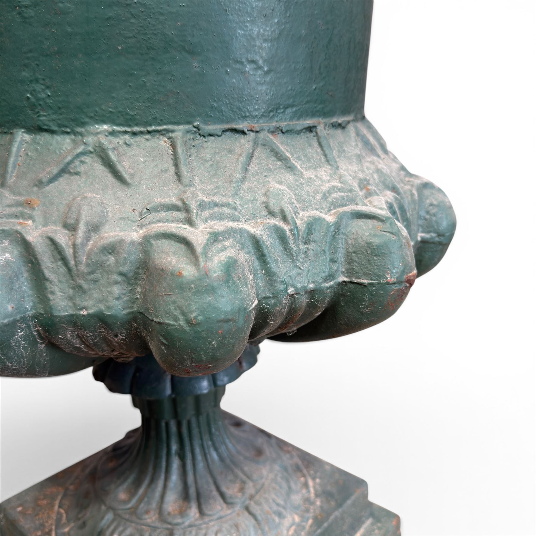 Pair of Victorian cast iron campana garden urns, the flared rim with repeating stylised anthemion motifs with scrollwork, over a deep gadrooned and Aesthetic moulded underbelly, fluted and foliate lower bowls on square pedestal bases, in antique green-painted finish