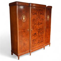 Early 20th century French burr maple breakfront armoire, enclosed by three panelled doors with bookmatched veneer and stringing, outer doors inlaid with gilt urn medallions, fitted interior with hanging spaces flanking a shelved compartment, raised on short tapered legs