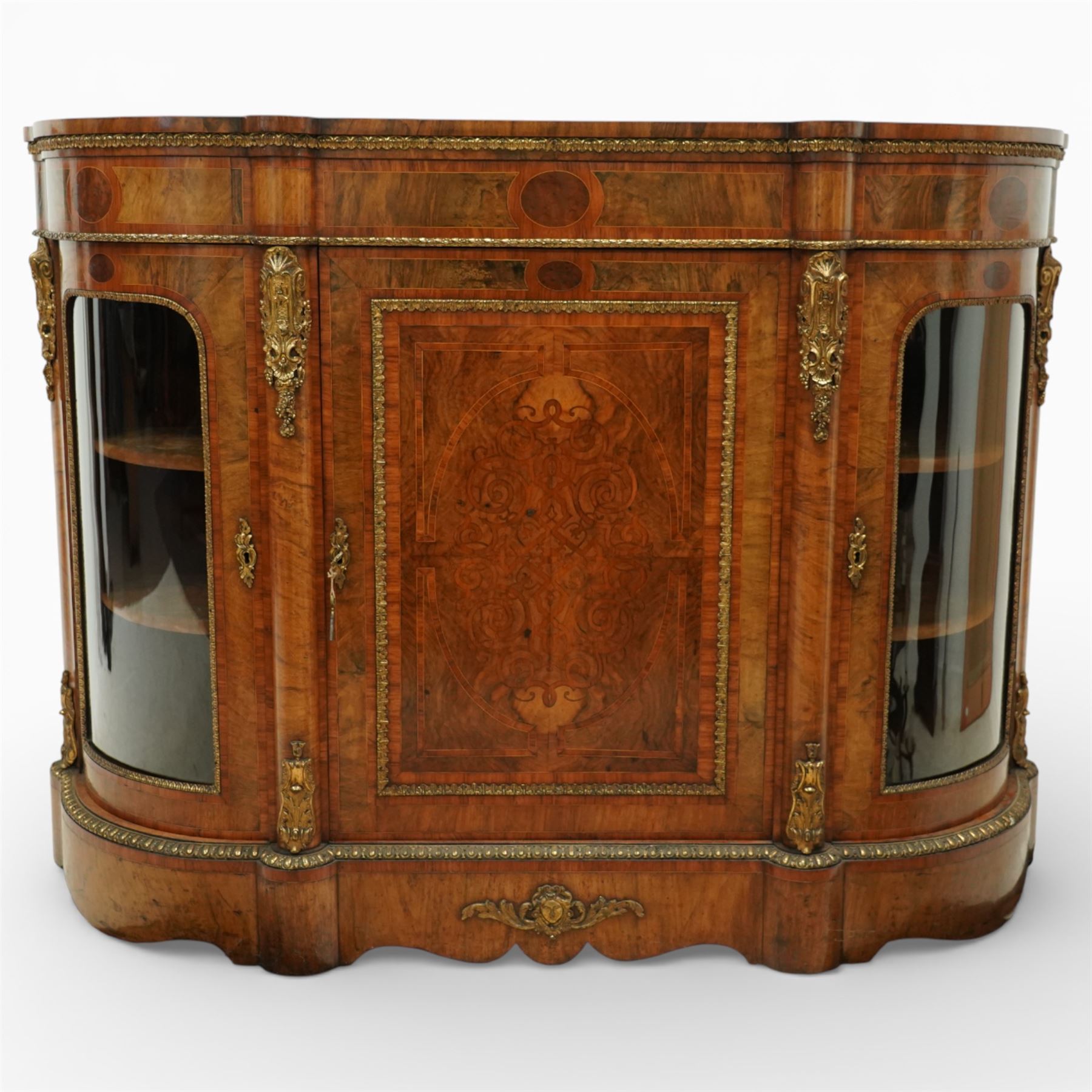 Late Victorian figured walnut credenza, the shaped demi-lune top with cross-banding and stringing over an inlaid frieze with foliate gilt-metal mounts, fitted with a central geometric-inlaid panelled door, flanked by convex-glazed side-cabinets with a gilt metal slip and outer-banding, the uprights surmounted by gilt-metal cartouche mounts with cascading foliage details, raised on a shaped skirted base with central female-mask gilt mount