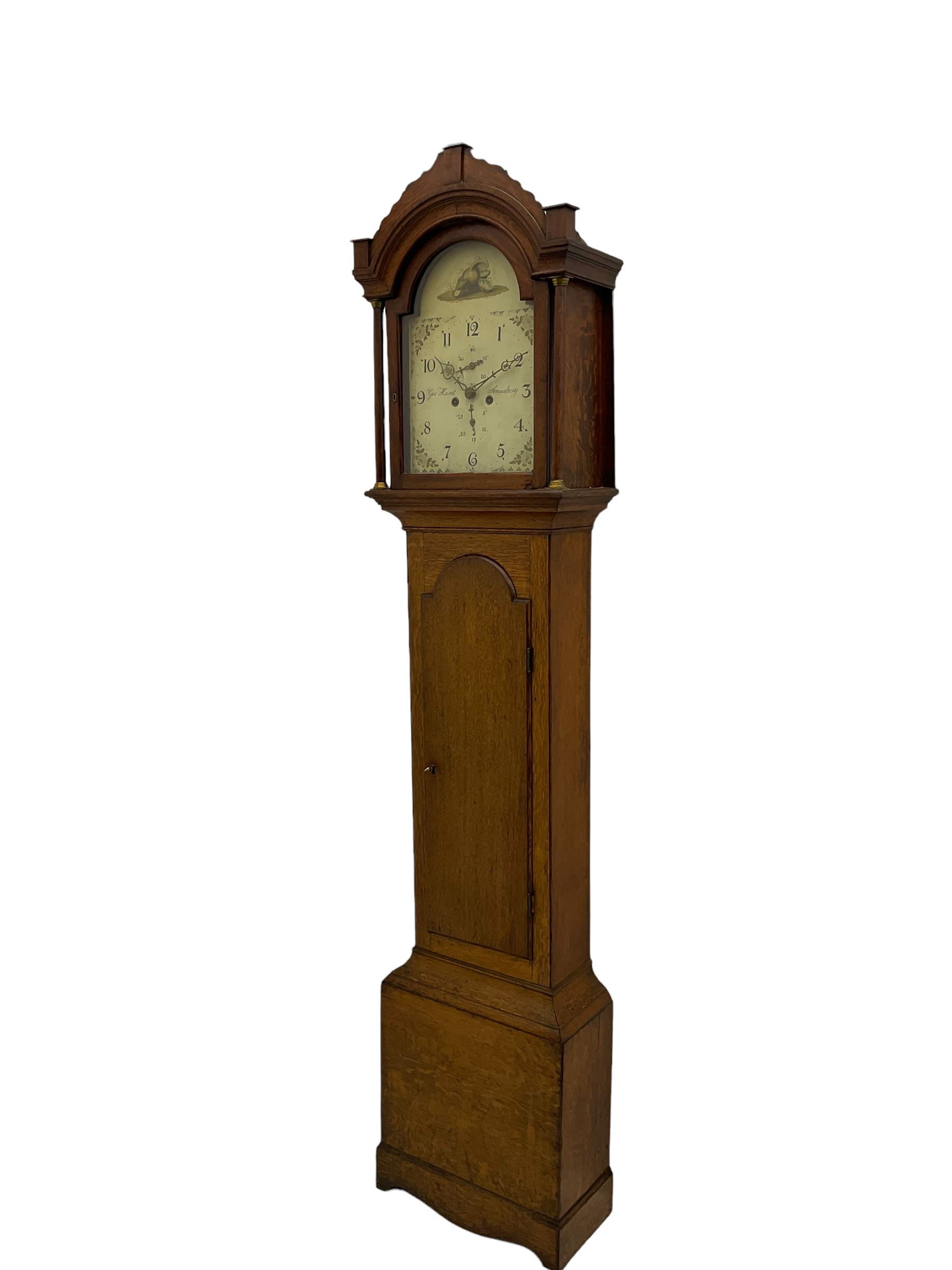 A provincial oak longcase clock retailed by “Geo Hunt, Amesbury” c1820, with a crested break arch pediment and corresponding glazed hood door flanked by two plain pillars with capitals, long trunk with a full length break arch door on a rectangular plinth with a shaped base, painted dial with Arabic numerals, minute markers, subsidiary date and seconds dials and stamped brass hands, matching painted spandrels and a depiction of sea shells to the arch, dial pinned to the movement via a Walker Hughes cast falseplate, with an eight day rack striking movement, striking the hours on a cast bell. With weights and Pendulum




