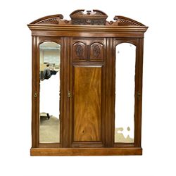 Victorian mahogany triple wardrobe, the pediment relief carved with urn and linen swags, the central panelled door carved with two fan motifs enclosing slides and drawers, two mirror glazed doors with bevelled plates enclosing hangings space and drawers, plinth base