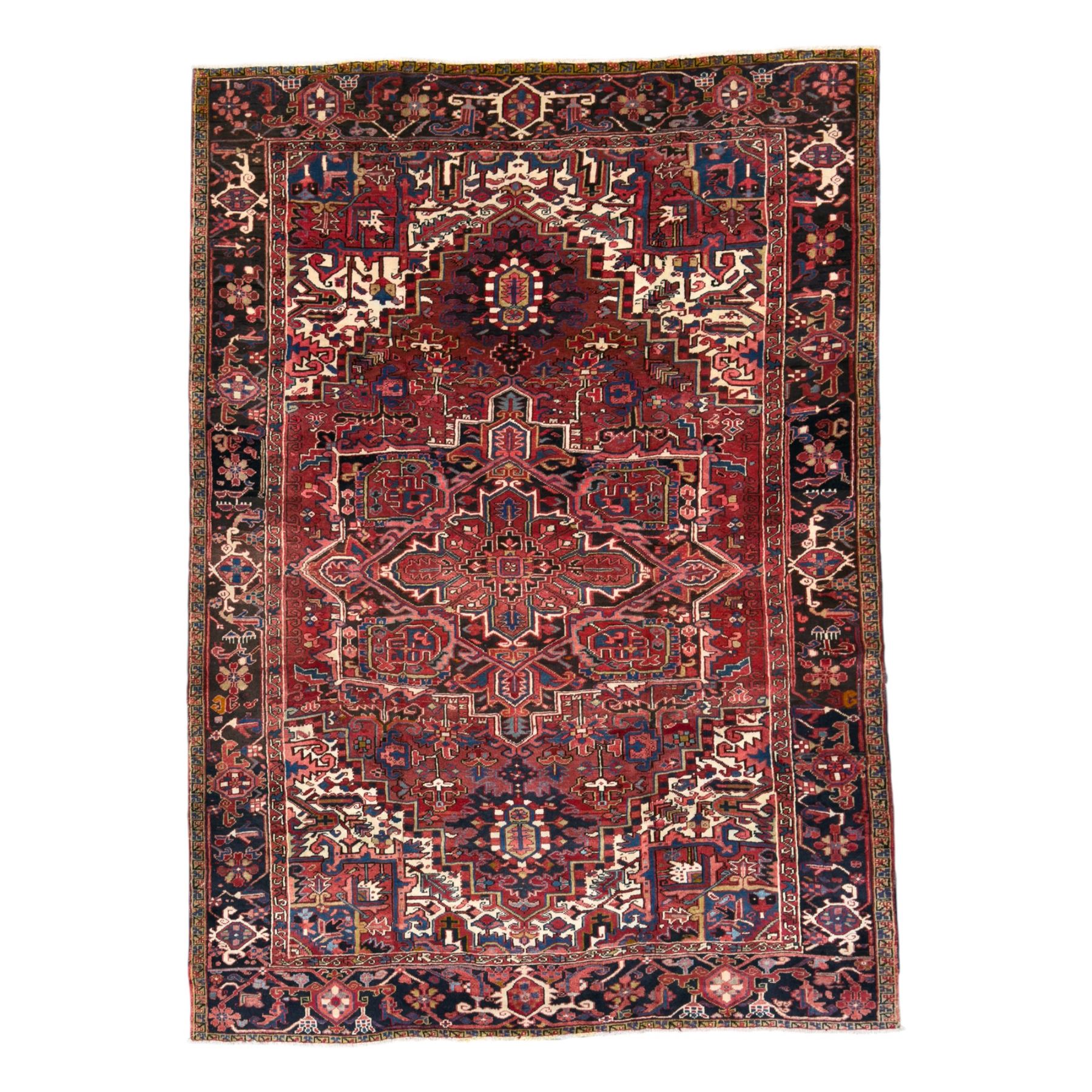 Northwestern Persian Heriz hand-knotted carpet, red madder field with a bold stepped medallion in indigo, ivory, and terracotta, flanked by angular floral motifs and stylized leaves, the spandrels set within a dark indigo border of rosettes and angular vine motifs between narrow guard stripes, 340cm x 245cm 