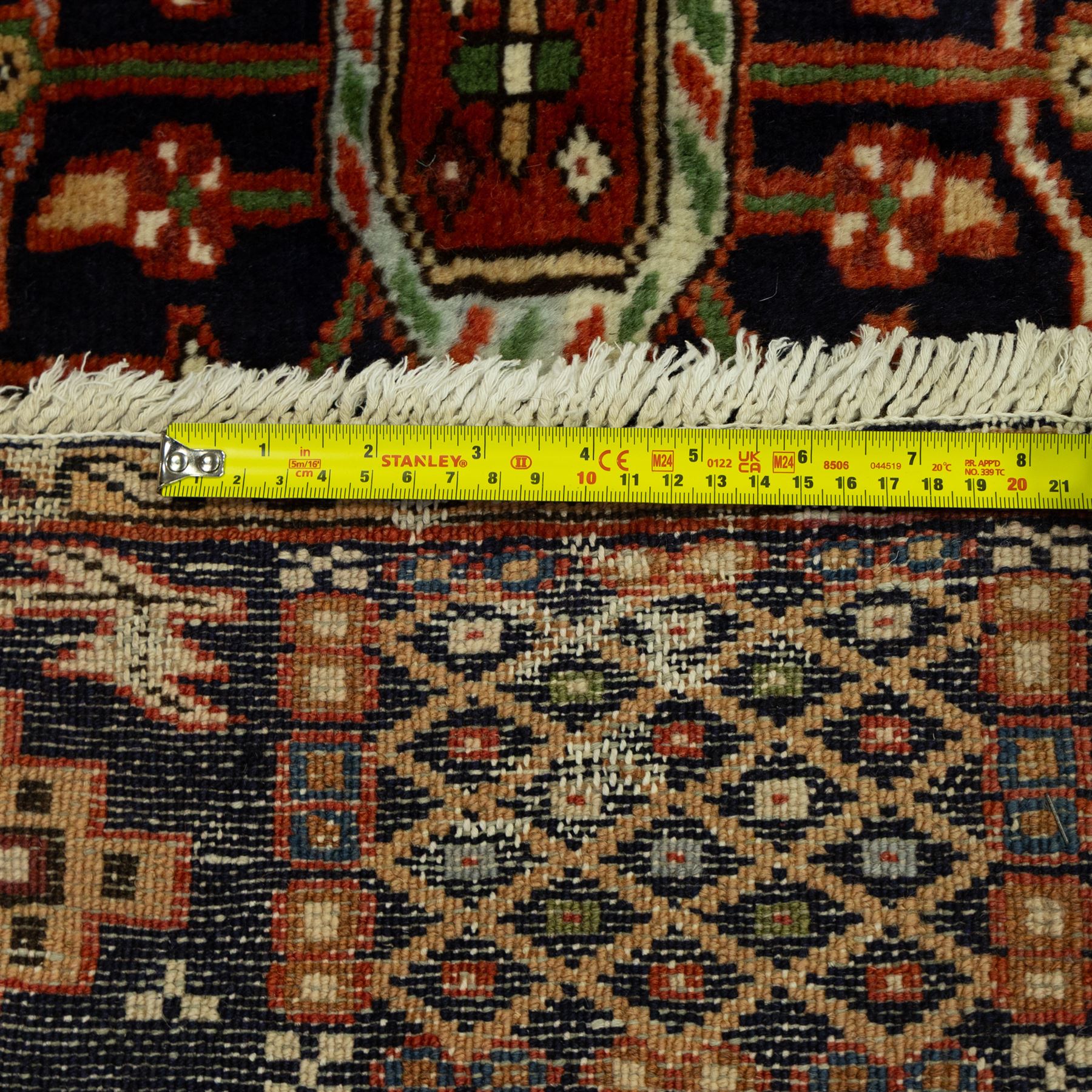 North West Persian Karajeh hand-knotted rug, terracotta ground, with a stepped geometric pole medallion design in blue, ivory and dark navy tones, surrounded by angular tribal motifs incorporating stylised flowerheads, hooked devices and geometric vine work, enclosed by a dark navy border with repeating rosettes and angular leaf motifs, flanked by narrow patterned guard stripes, with cotton fringes