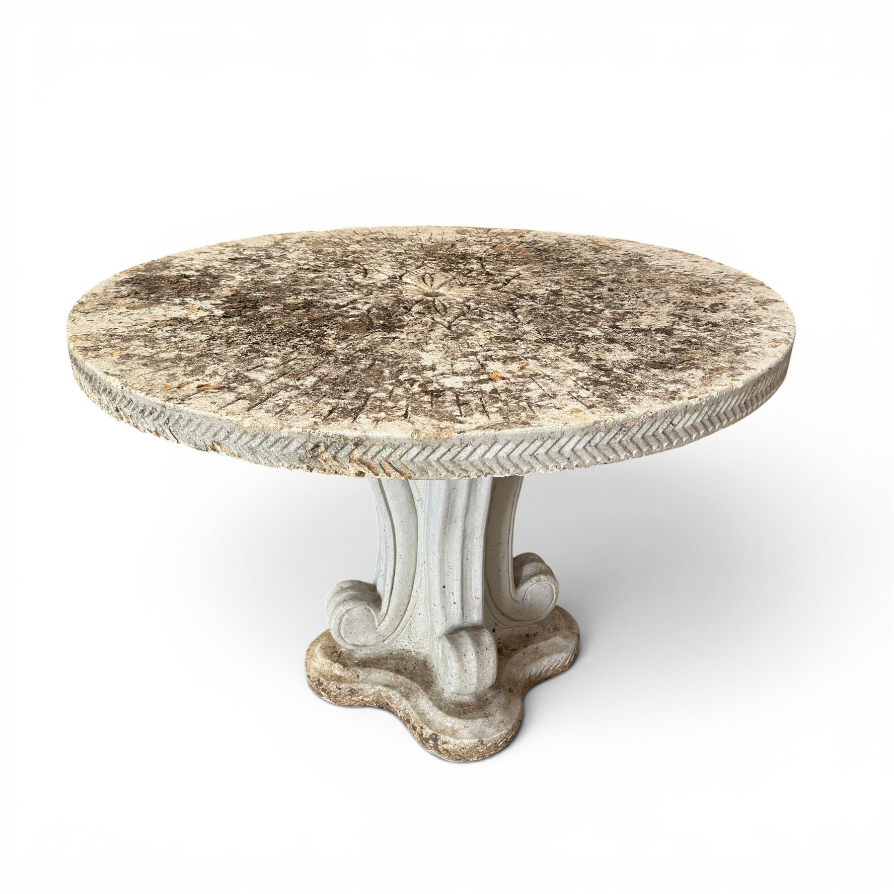 Weathered cast stone garden table, circular top etched with central floral medallion, the edge with repeating chevron motifs, raised on a quadriform S-scroll pedestal with stepped and chevron carved base