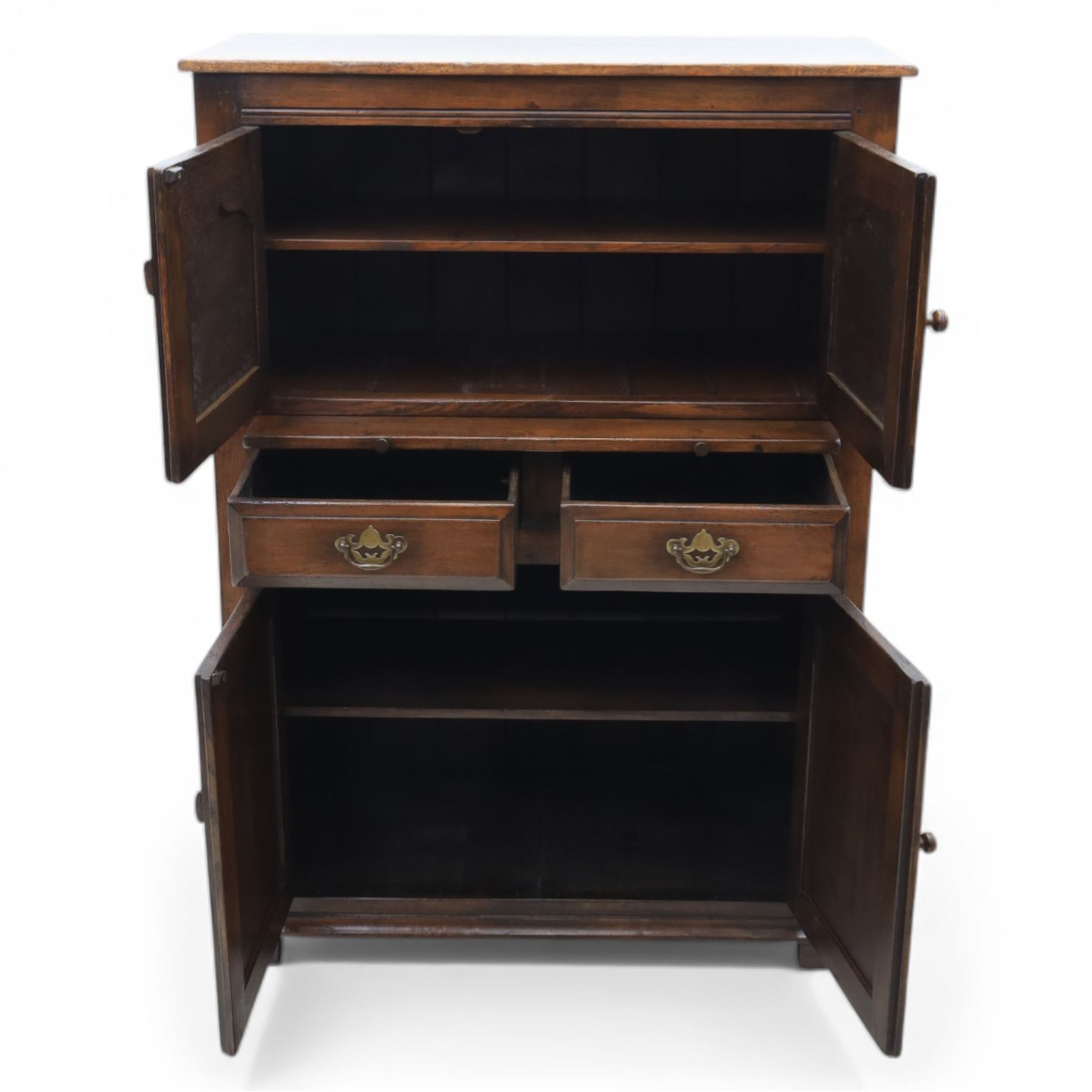  Late 20th century oak four door cupboard in the manner of Titchmarsh and Goodwin, rectangular plank top above two pairs of fielded panel doors with shaped raised panels, central arrangement of a brushing slide above two short drawers with brass swan neck handles and pierced backplates, enclosed by further pair of panelled cupboard doors, fitted with brass butt hinges and turned wooden knobs, raised on plinth base