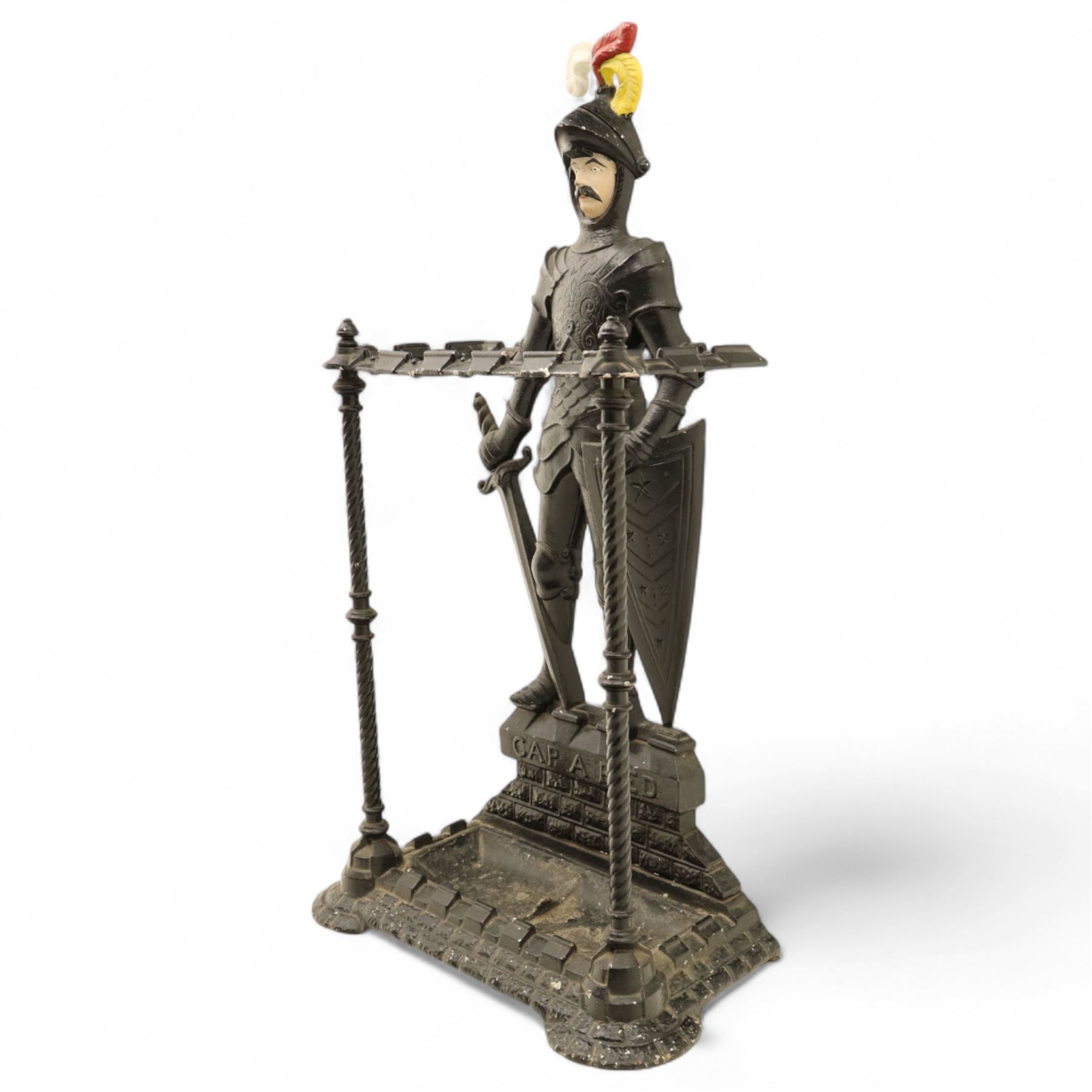 Early 20th century painted cast iron hall stand, formed as a standing armoured figure with red and yellow feathered crest, twin rails and integral drip tray with castellated edge